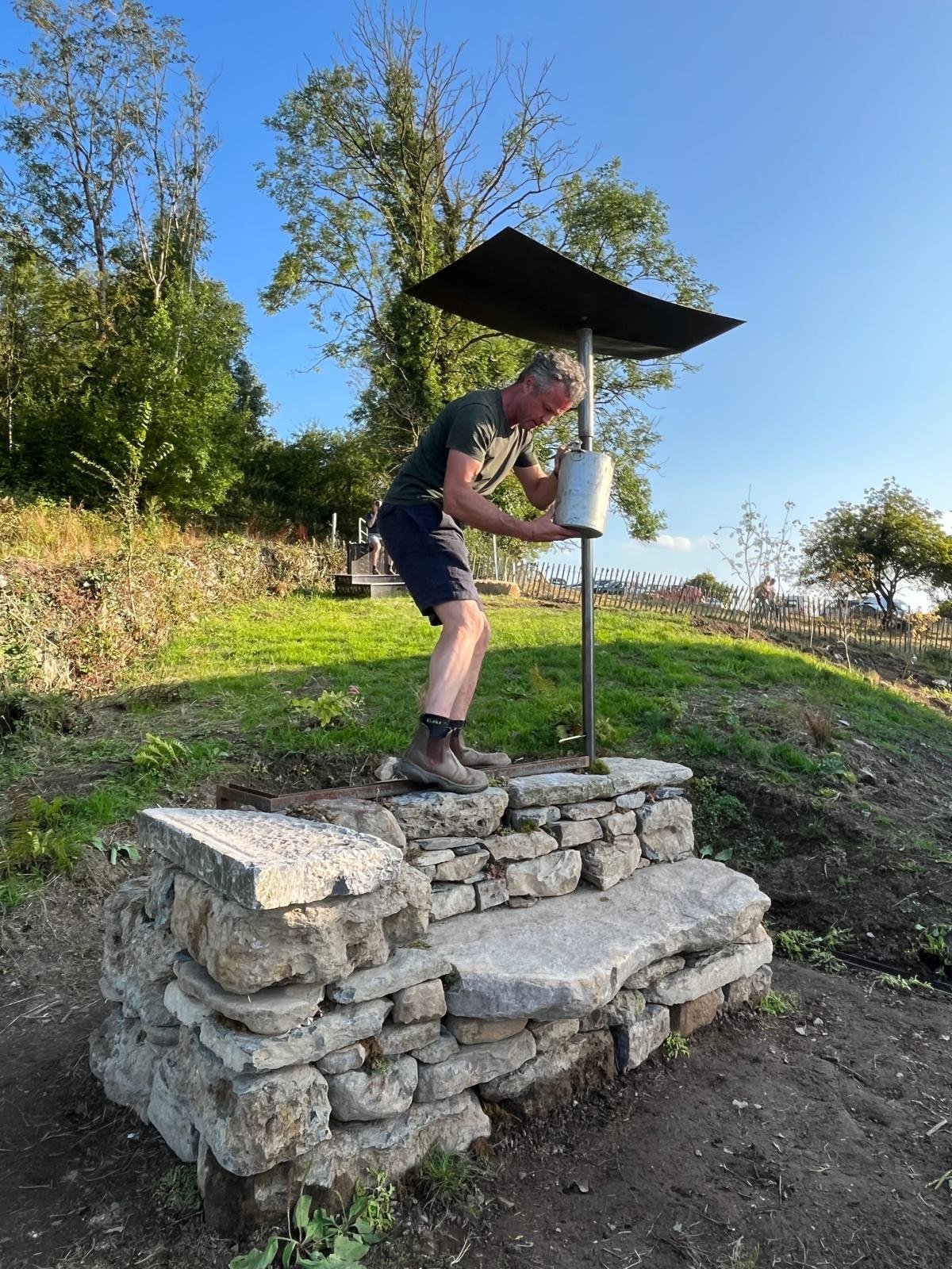 A man standing on a stone structure outdoors, examining a water pump with a shade above it, in a grassy area with trees and a clear blue sky.