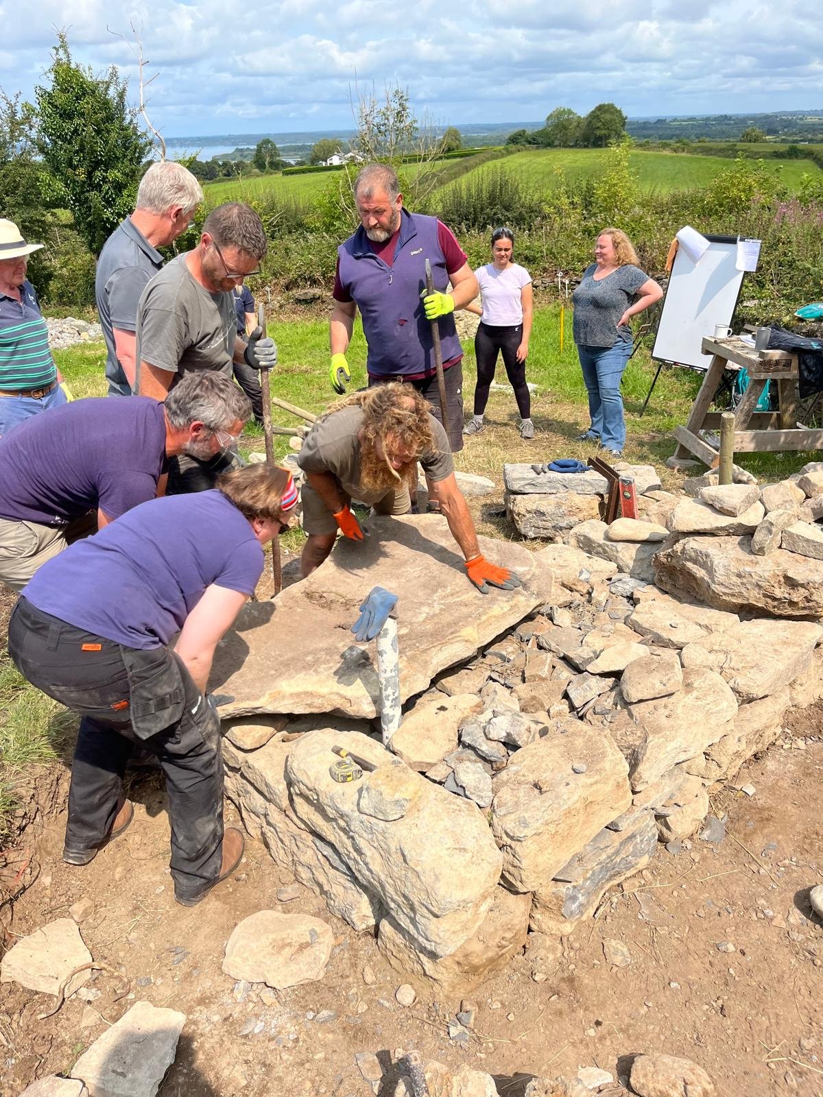 A group of people working outdoors on a stone construction project, with some individuals observing and others actively handling large rocks and tools, in a rural landscape with green fields and a cloudy sky.