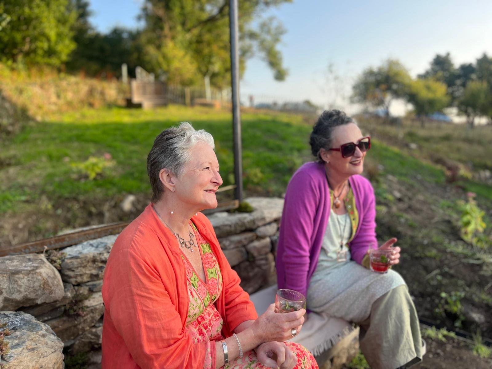 Two elderly women sitting outdoors on a stone bench, enjoying drinks and smiling while talking, with a grassy hill and trees in the background during daytime.