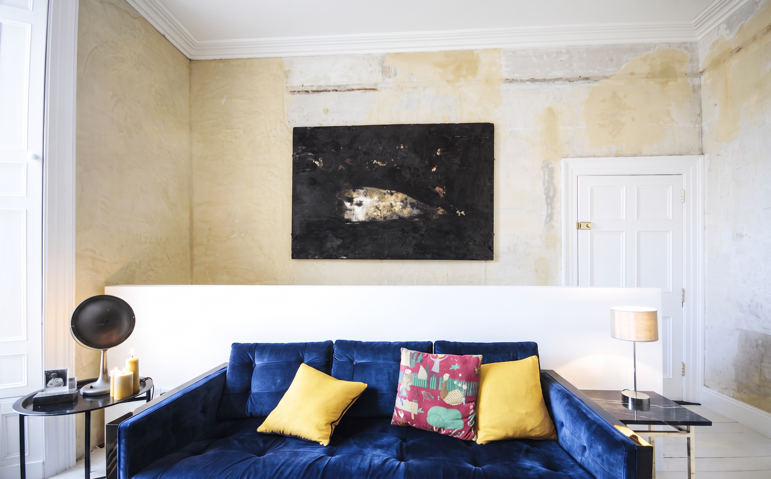 Living room with a dark blue velvet sofa with yellow and colorful patterned pillows, a black side table with candles and a photo frame, a white wall with an abstract black painting, and a white door with decorative molding.