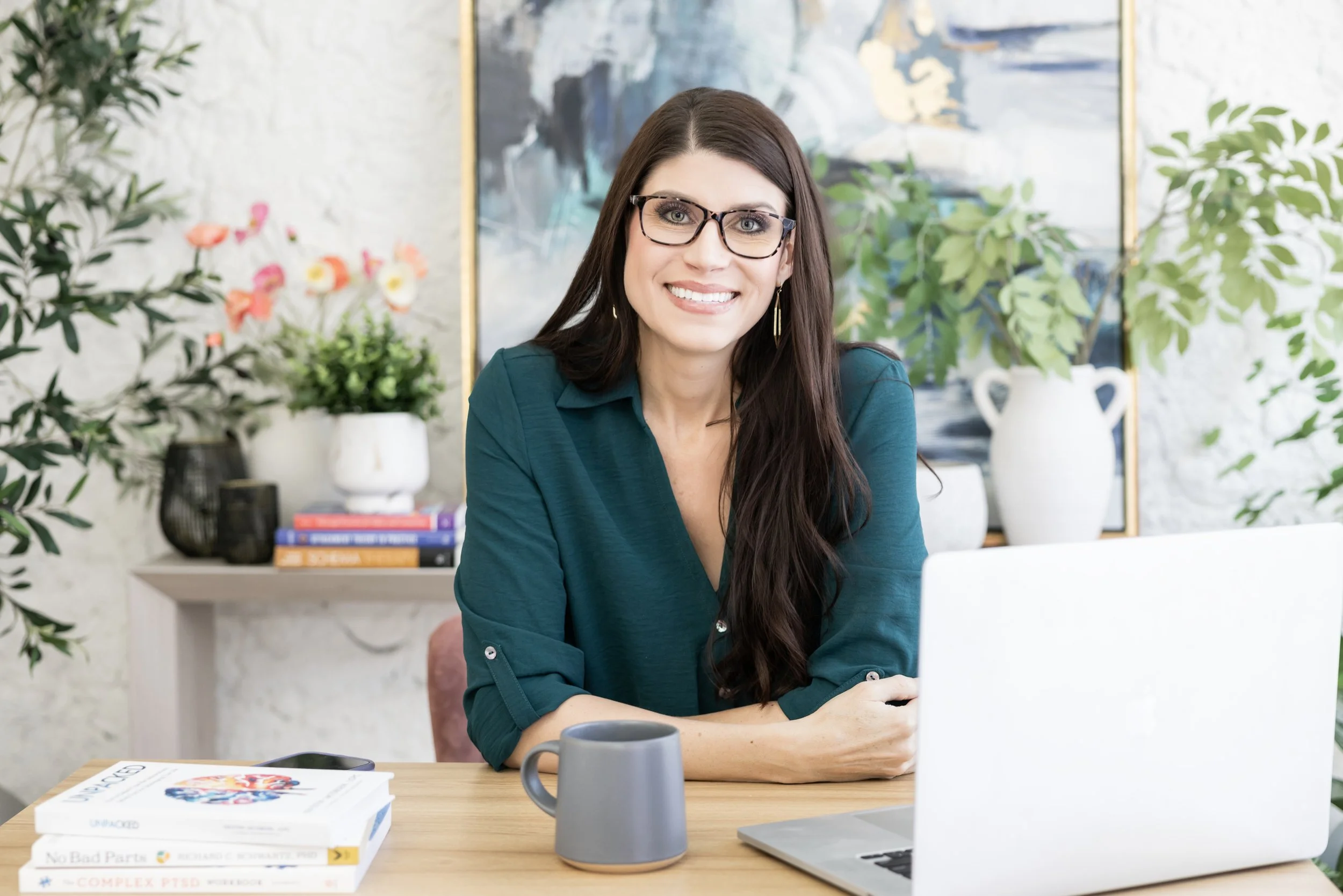 A woman with long dark hair and glasses sitting at a desk in a well-decorated office or home workspace, smiling at the camera with a laptop, books, a coffee mug, and plants around her.