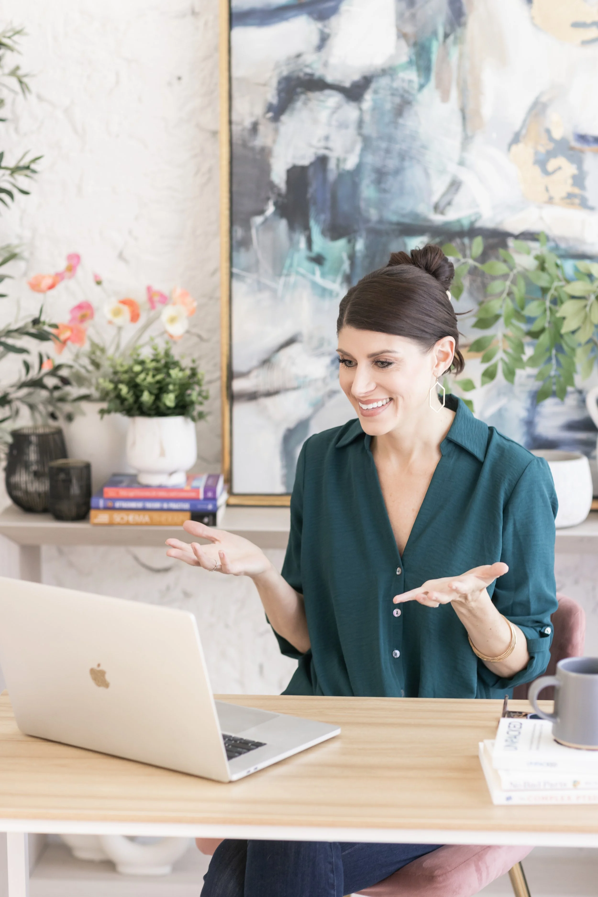A woman in a teal shirt smiling and gesturing with her hands while sitting at a desk with a laptop, a mug, and books, in a bright room with a large abstract painting and potted plants.