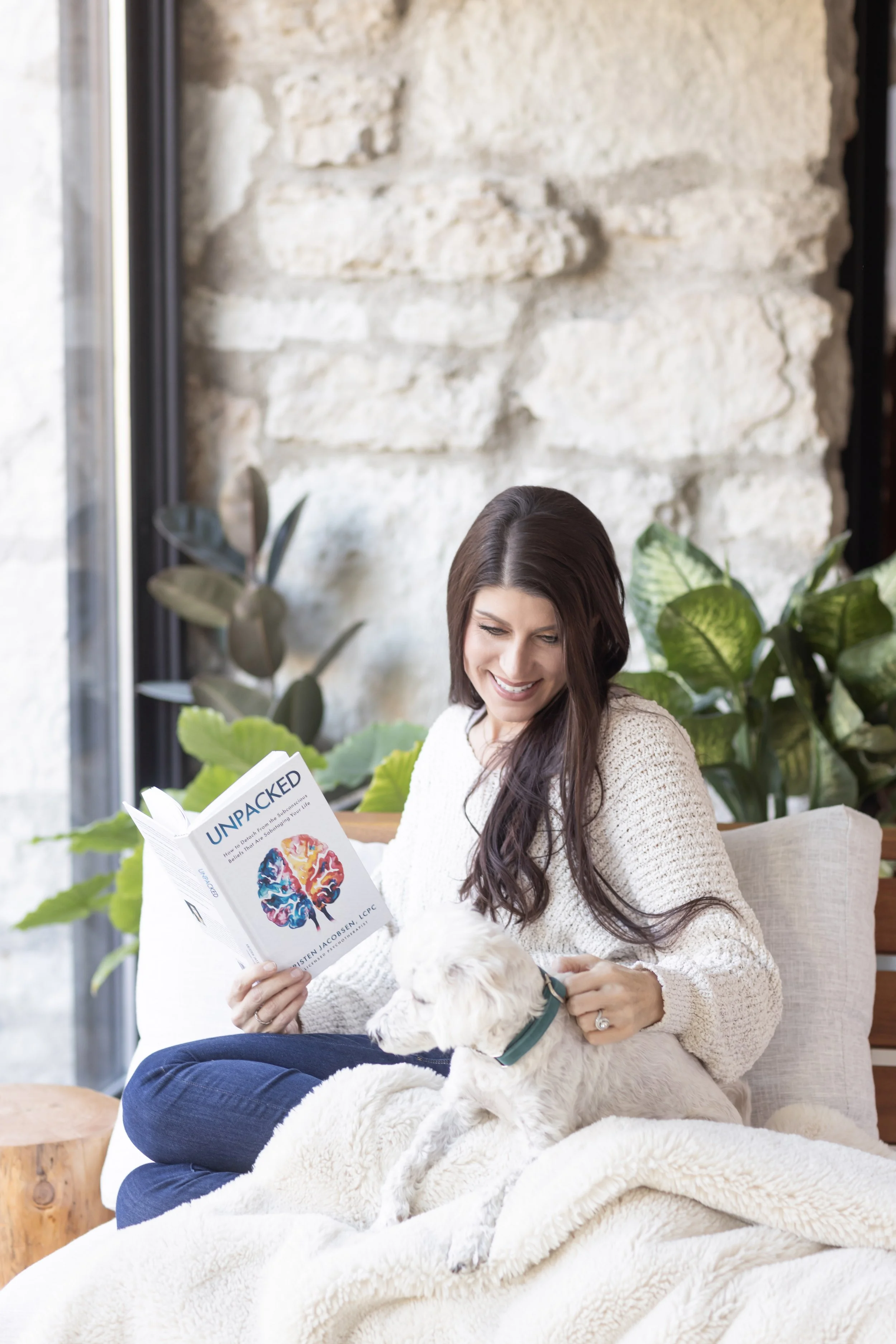 Woman sitting on a beige sofa with a white dog, reading a book titled 'Unpacked' inside a cozy room with large windows, greenery, and a stone wall.