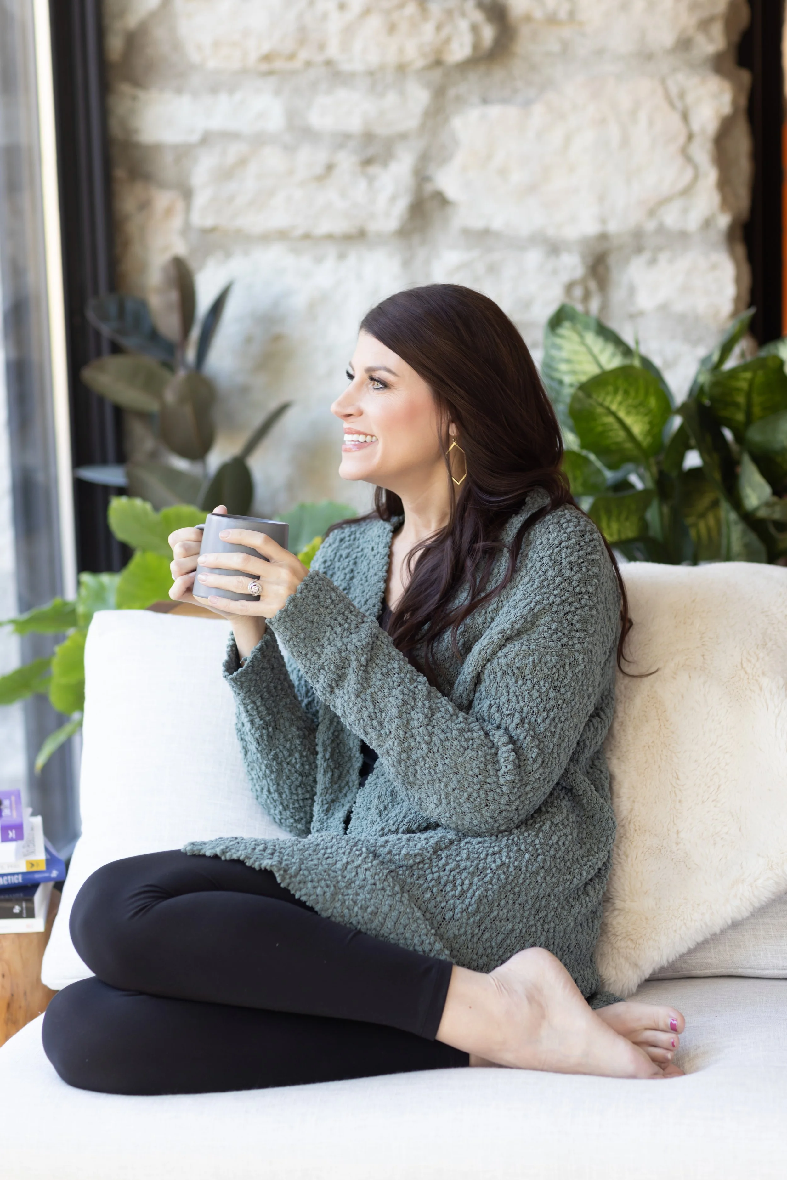 A woman with long dark hair smiling and holding a gray mug while sitting on a beige sofa with a fluffy pillow, surrounded by green plants and a stone wall.