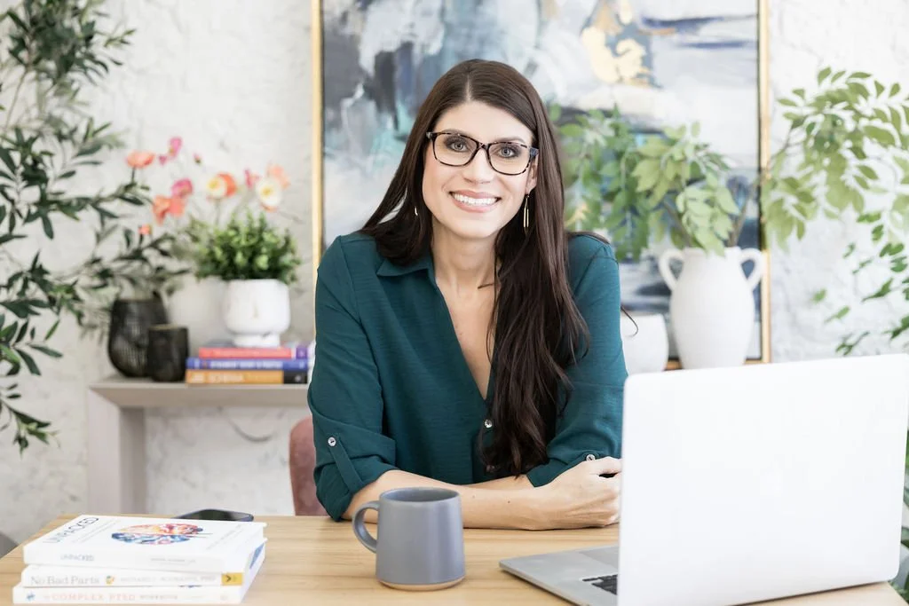A woman with long dark hair, glasses, and a teal blouse sitting at a desk with a laptop, coffee mug, and books, smiling in a bright, decorated room.