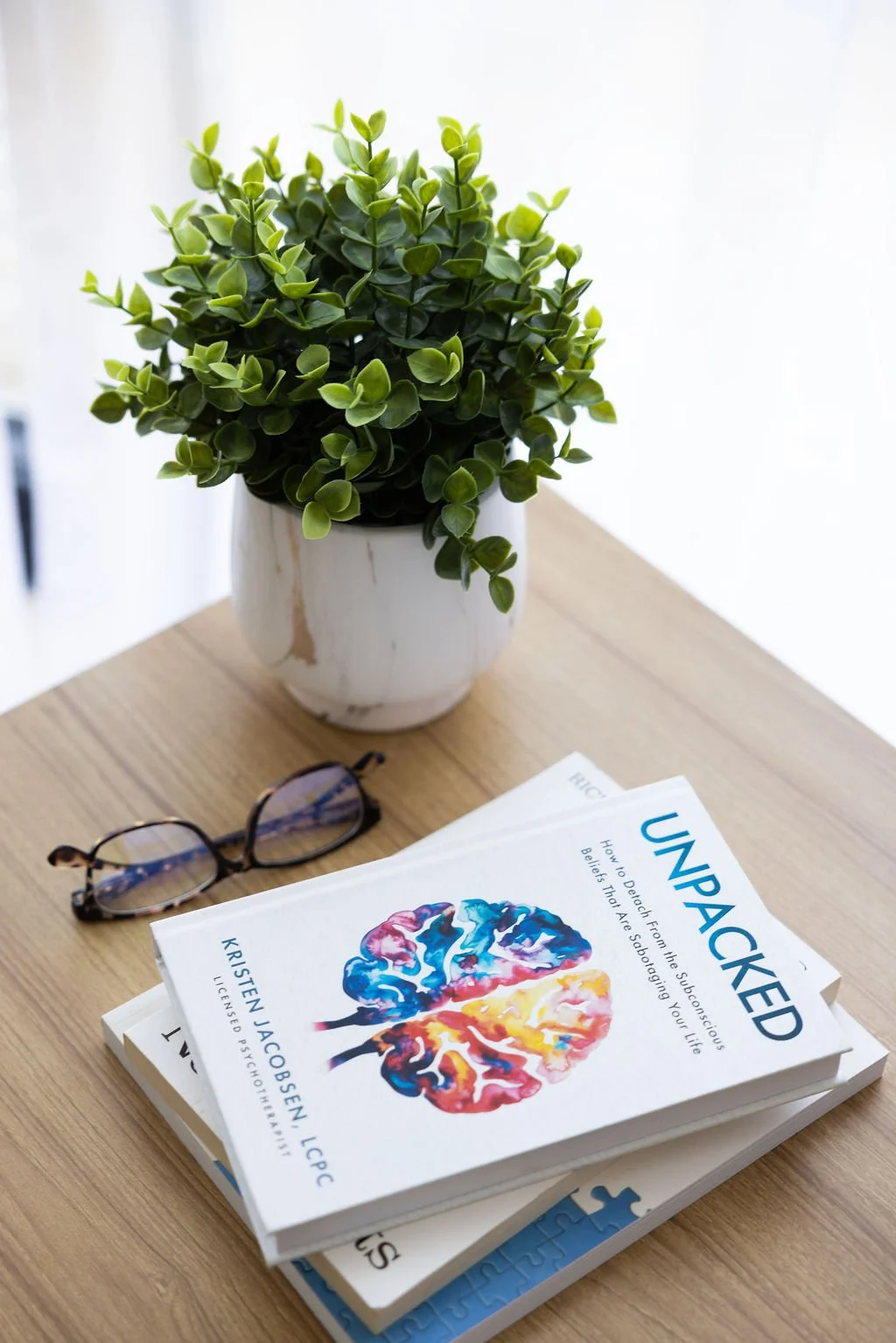 A potted green plant, a pair of glasses, and a stack of books on a wooden table.