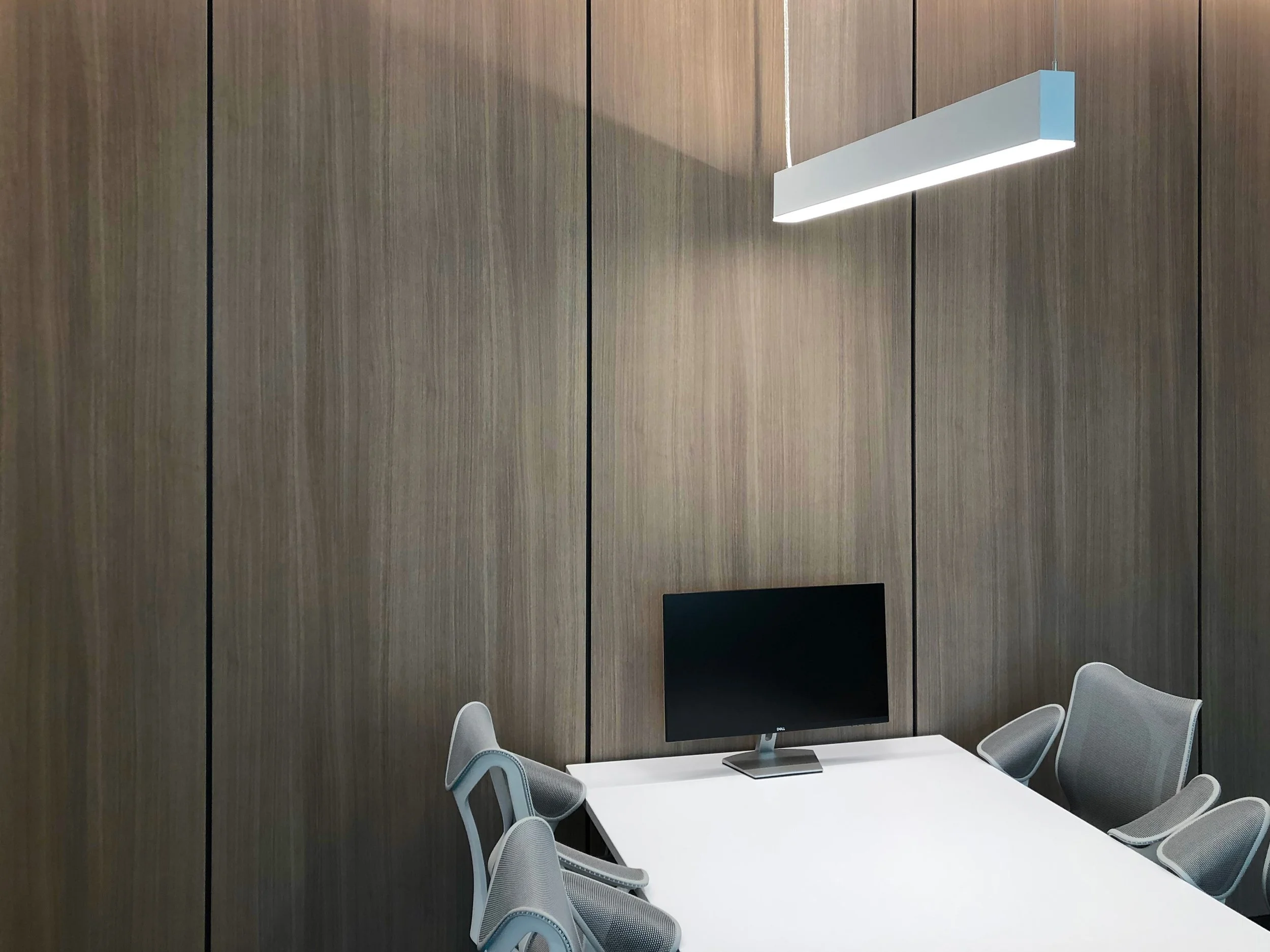 Modern conference room with wooden panel walls, a white table, gray mesh chairs, and a large flat-screen monitor on the wall.