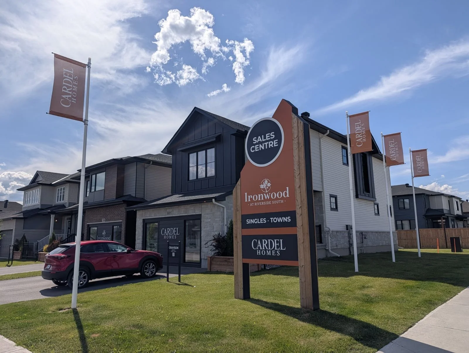 Exterior view of a row of modern houses with a sales center sign in front. The sign reads 'Sales Centre' for Ironwood at Riverside South by Cardel Homes. Flags with 'Cardel Homes' are flying in the sky. A red vehicle is parked on the street on a sunny day with a partly cloudy sky.