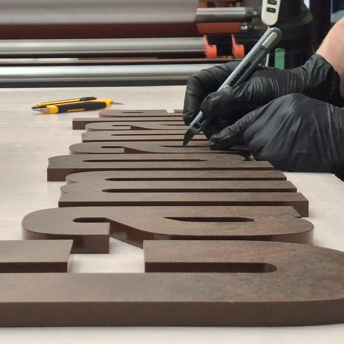 Person wearing black gloves working on a large brown wooden letter on a worktable in a workshop.