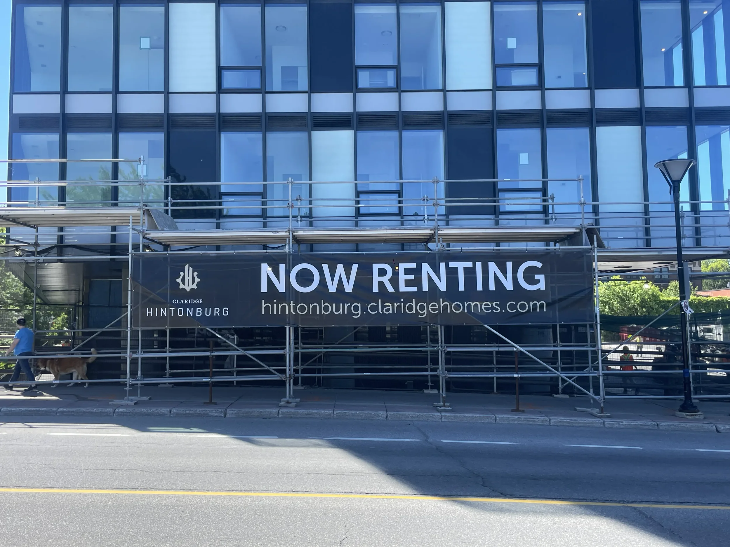 Building with glass windows under construction or renovation, scaffolding in front, and a black sign that says 'NOW RENTING' with contact information.