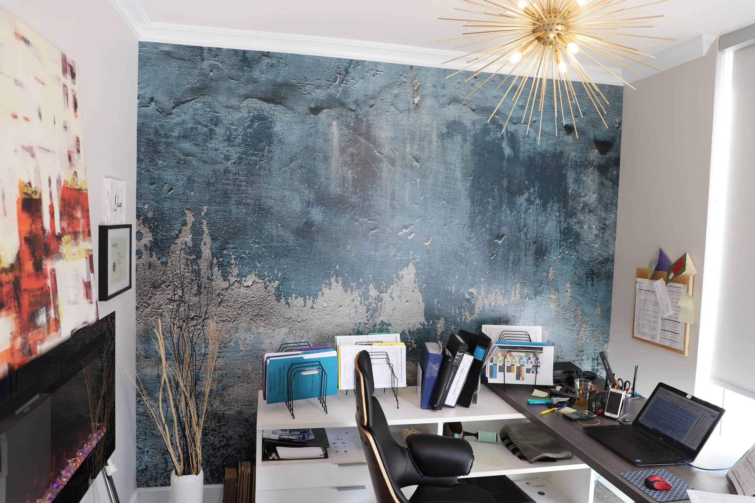 Home office with a black desk, white shelves, computer, laptop, water glass, binders, office supplies, art on a textured blue wall, a gold starburst chandelier, and a corner of a white wall with a corkboard.