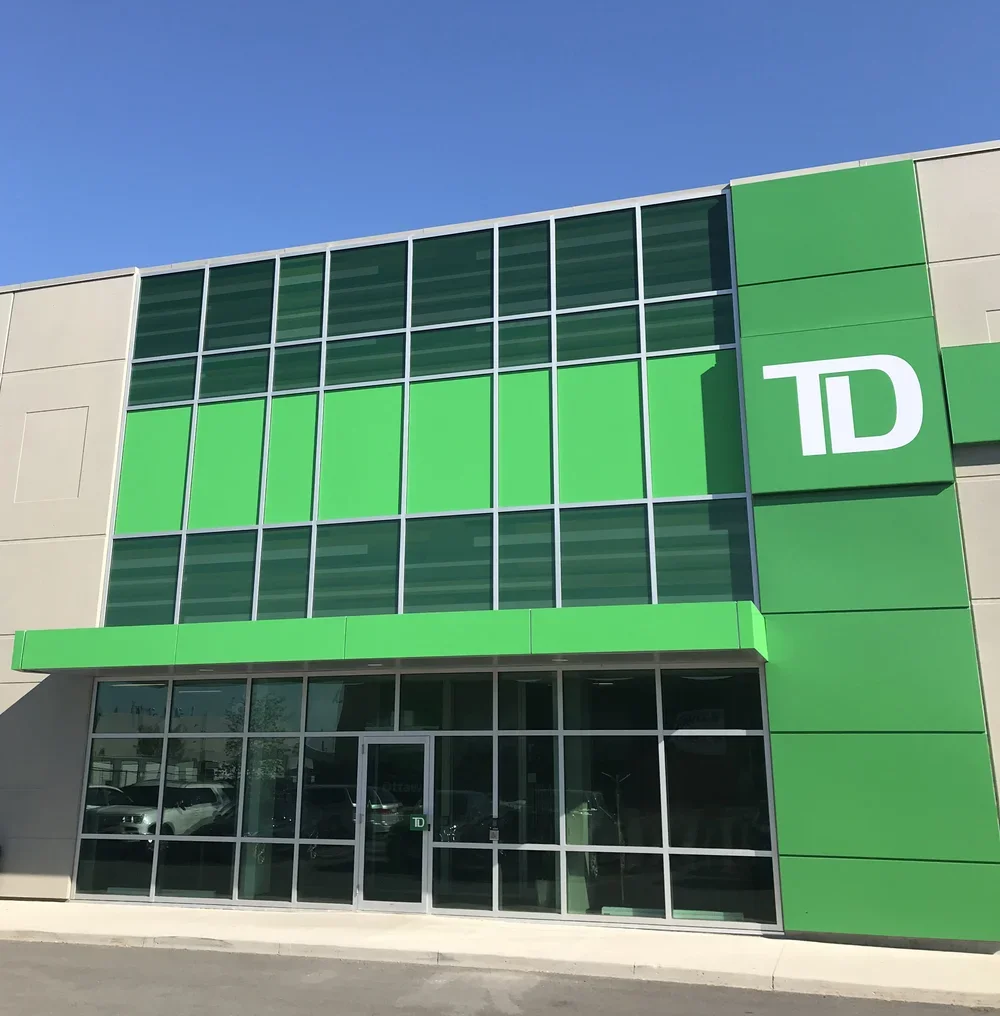 A modern commercial building with a green and beige exterior, featuring large windows and a green door, with a prominent green sign displaying 'TD' on the front facade.
