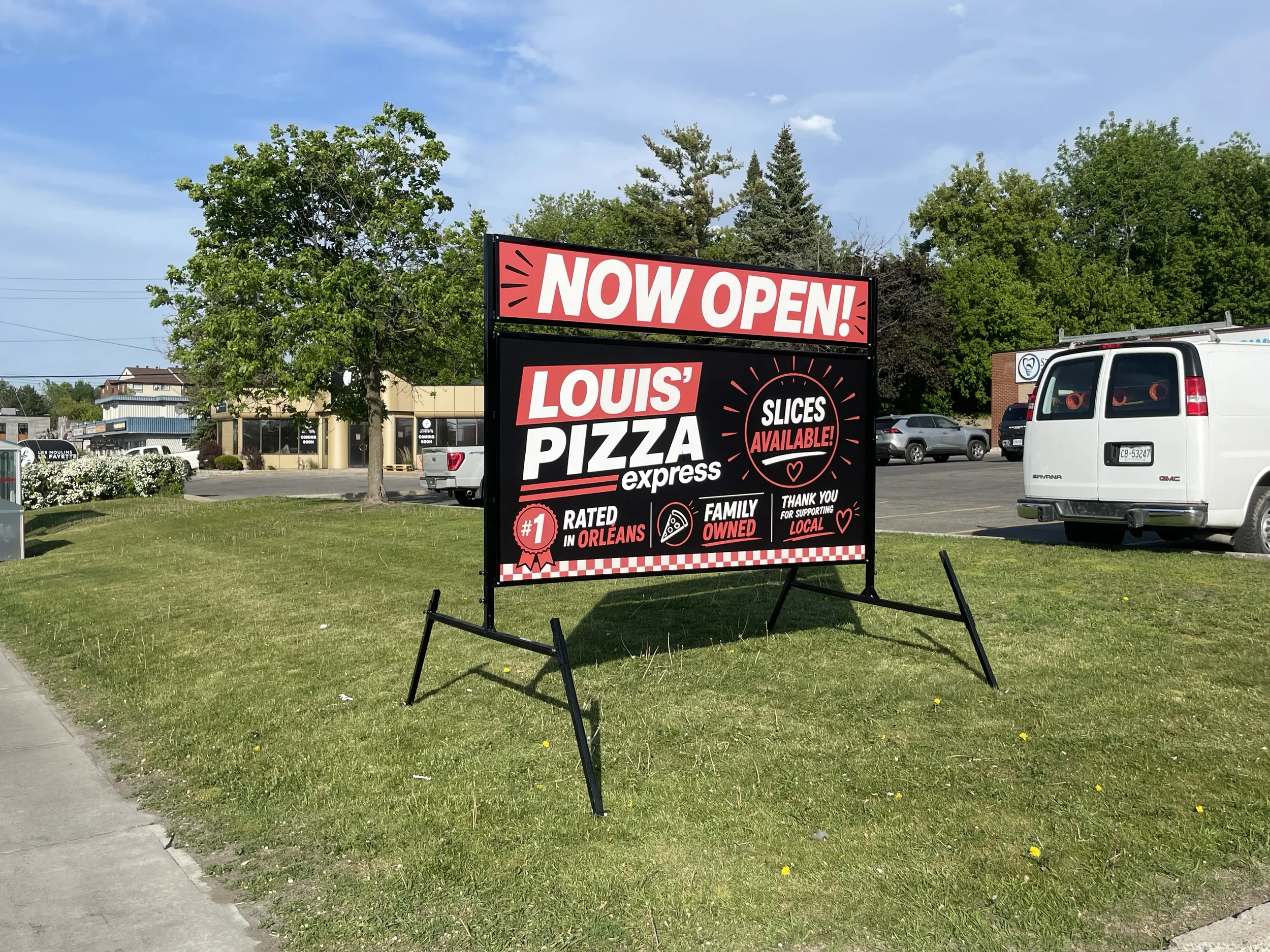 A large sign in a parking lot displays 'NOW OPEN! Louis' Pizza express' with additional text about slices being available, and highlights about being rated number 1 in Orleans, family owned, and supporting local businesses. The sign is set amongst green grass and trees, with cars, buildings, and a blue sky in the background.