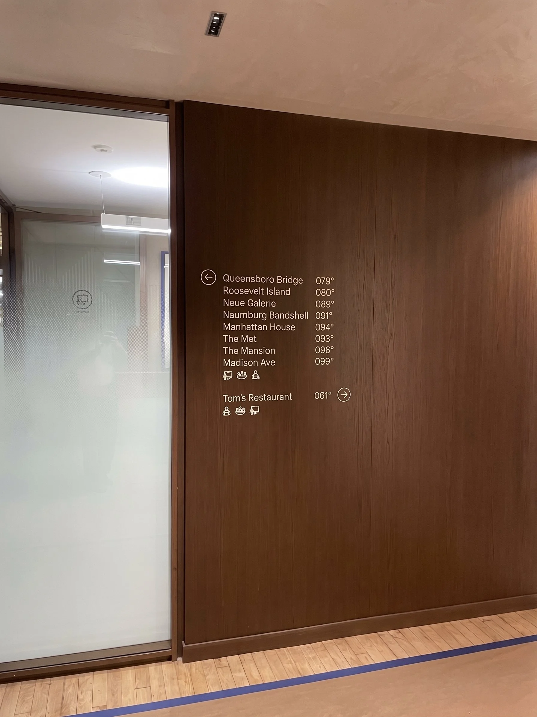 Indoor hallway with a wooden wall, a frosted glass door on the left, and a doorway with a sign displaying directions and nearby icons.