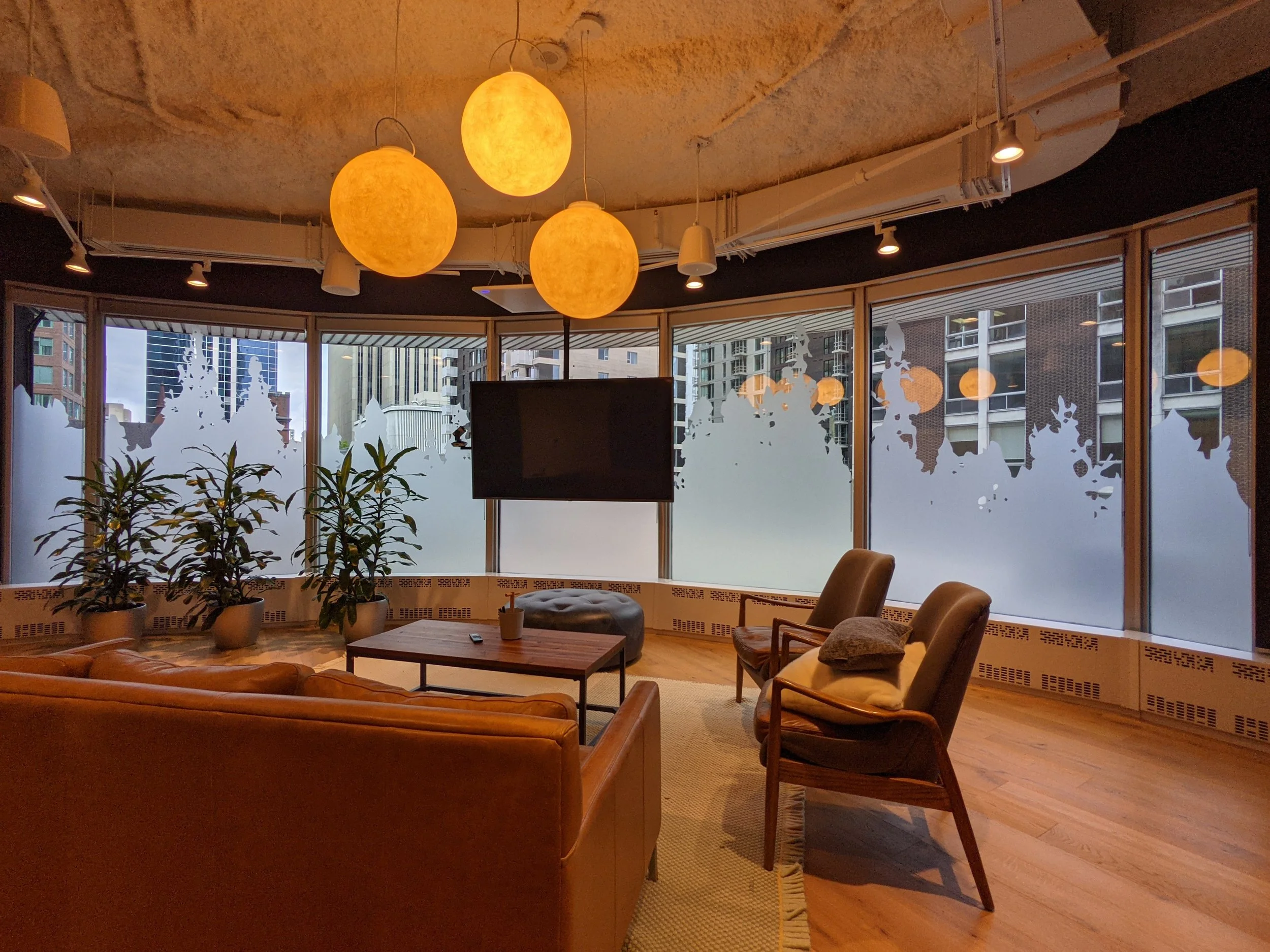 Modern office lounge with large windows with artistic window frosting, hanging spherical lamps, leather sofa, wooden chairs, a TV, and potted plants.