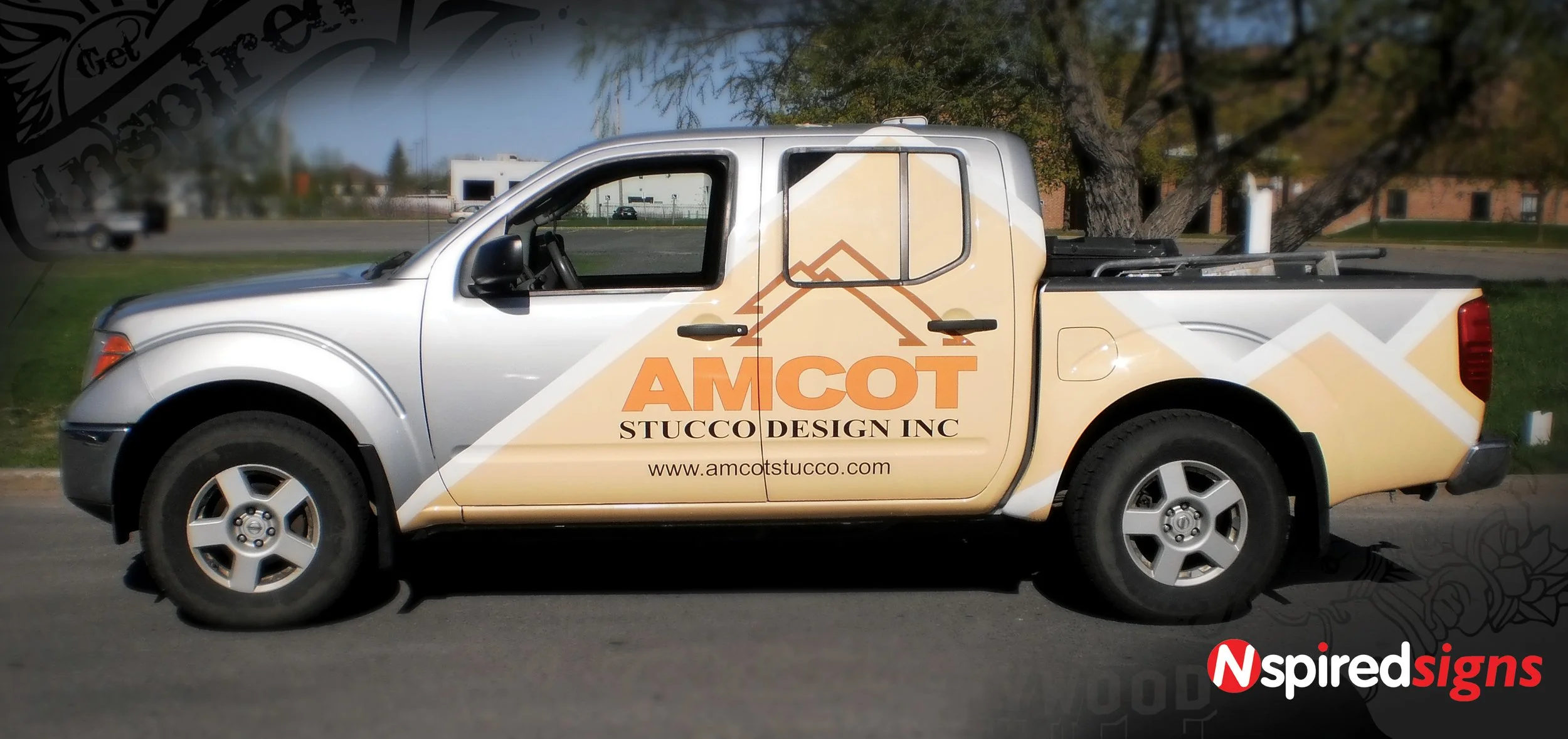 A silver pickup truck with a large graphic wrap advertising AMCOT Stucco Design Inc., featuring a mountain range logo and website www.amcotstucc.com, parked on a street with trees in the background.