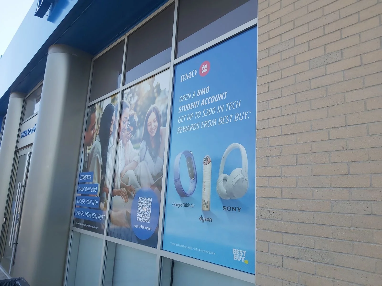 Exterior of a retail store with large advertisement poster promoting BMO student bank account and tech rewards, featuring images of a Fitbit Air, Dyson, and Sony headphones.