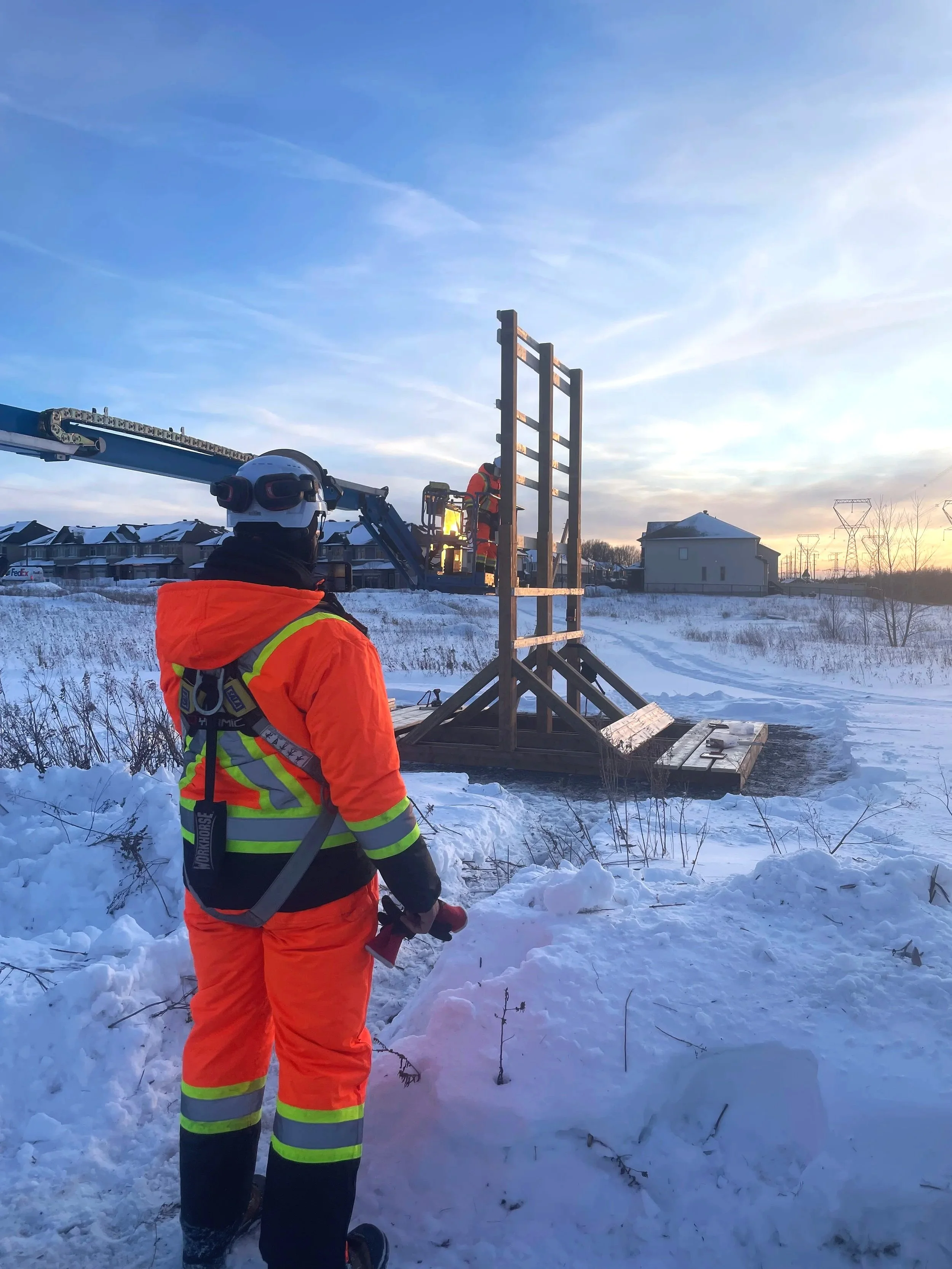 Emergency workers in orange and yellow safety gear setting up a tall observation or support structure in a snowy area at sunset.