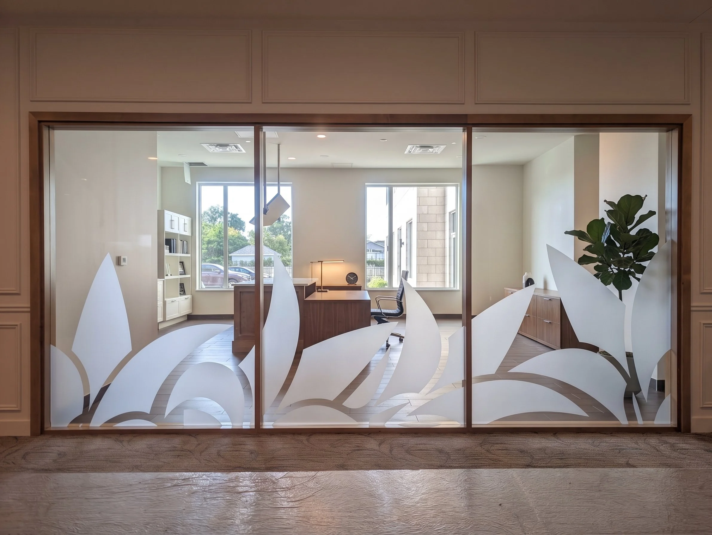 View through glass door with floral design on it into a modern office space with desk, chair, bookshelf, and large windows showing parked cars and trees outside.