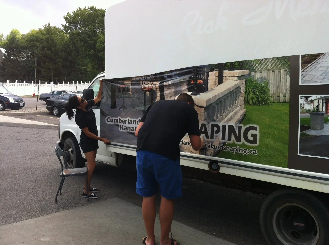 Two people applying a vinyl sign to the side of a white-moving truck, with a woman holding the sign and a man attaching it, advertising landscaping services.
