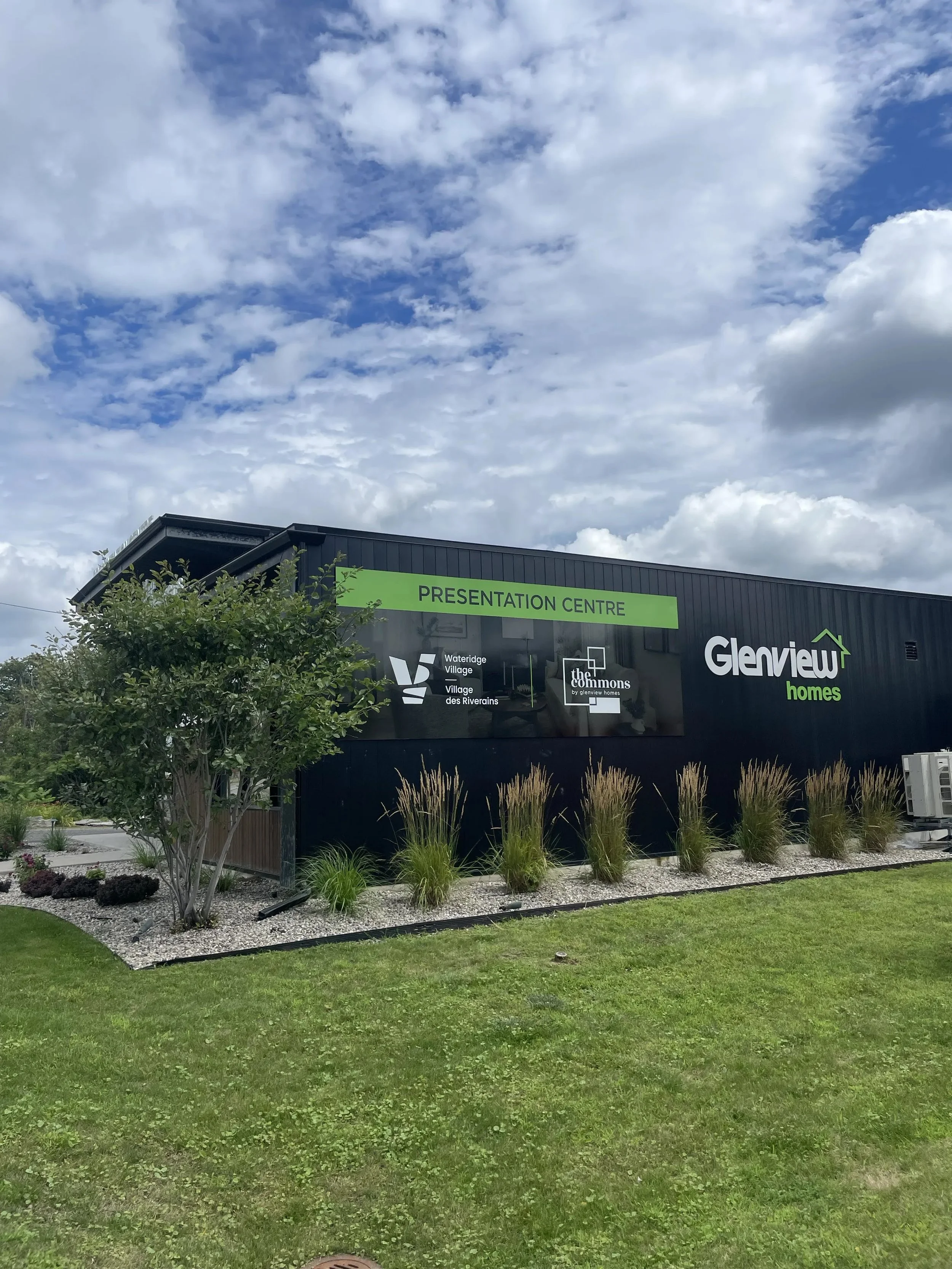A modern black building with green signage that reads 'Presentation Centre' and 'Glenview Homes', surrounded by landscaped greenery including a small tree, ornamental grass, and neatly maintained grass lawn, with a partly cloudy sky overhead.