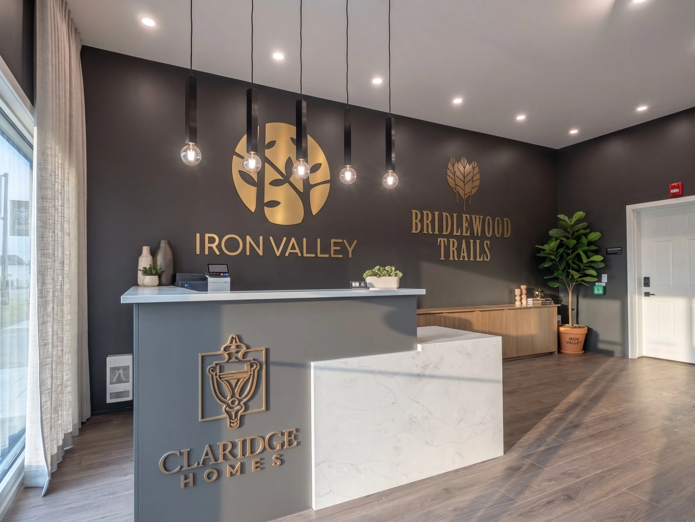 Interior of a home showroom with a dark gray wall, showcasing a logo with a stylized tree and the words 'Iron Valley,' 'Bridlewood Trails,' and 'Claredge Homes' in gold lettering. The reception desk in the foreground has decorative vases, a plant, and a marble top. There are recessed lights in the ceiling and hanging pendant lights over the desk. A potted plant is to the right near the door.