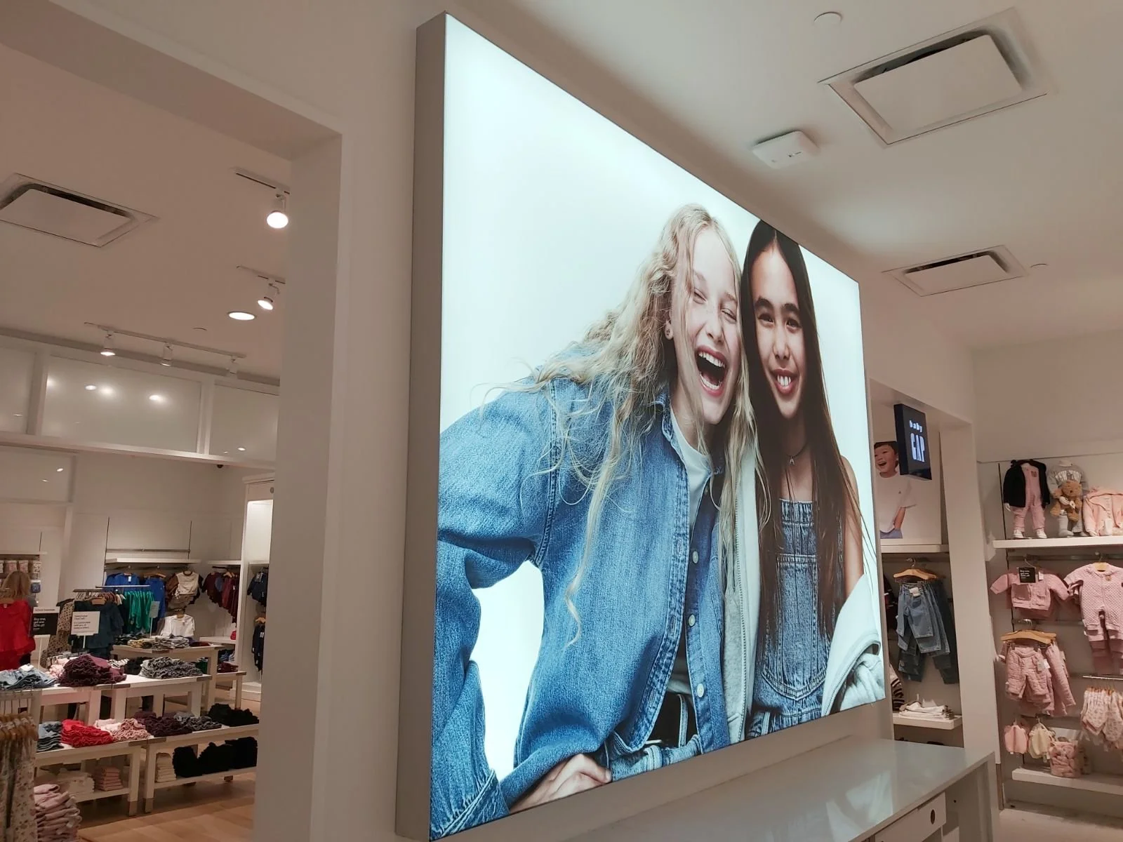 A large advertisement display in a retail store shows two young women smiling and laughing, wearing denim jackets and casual clothing.