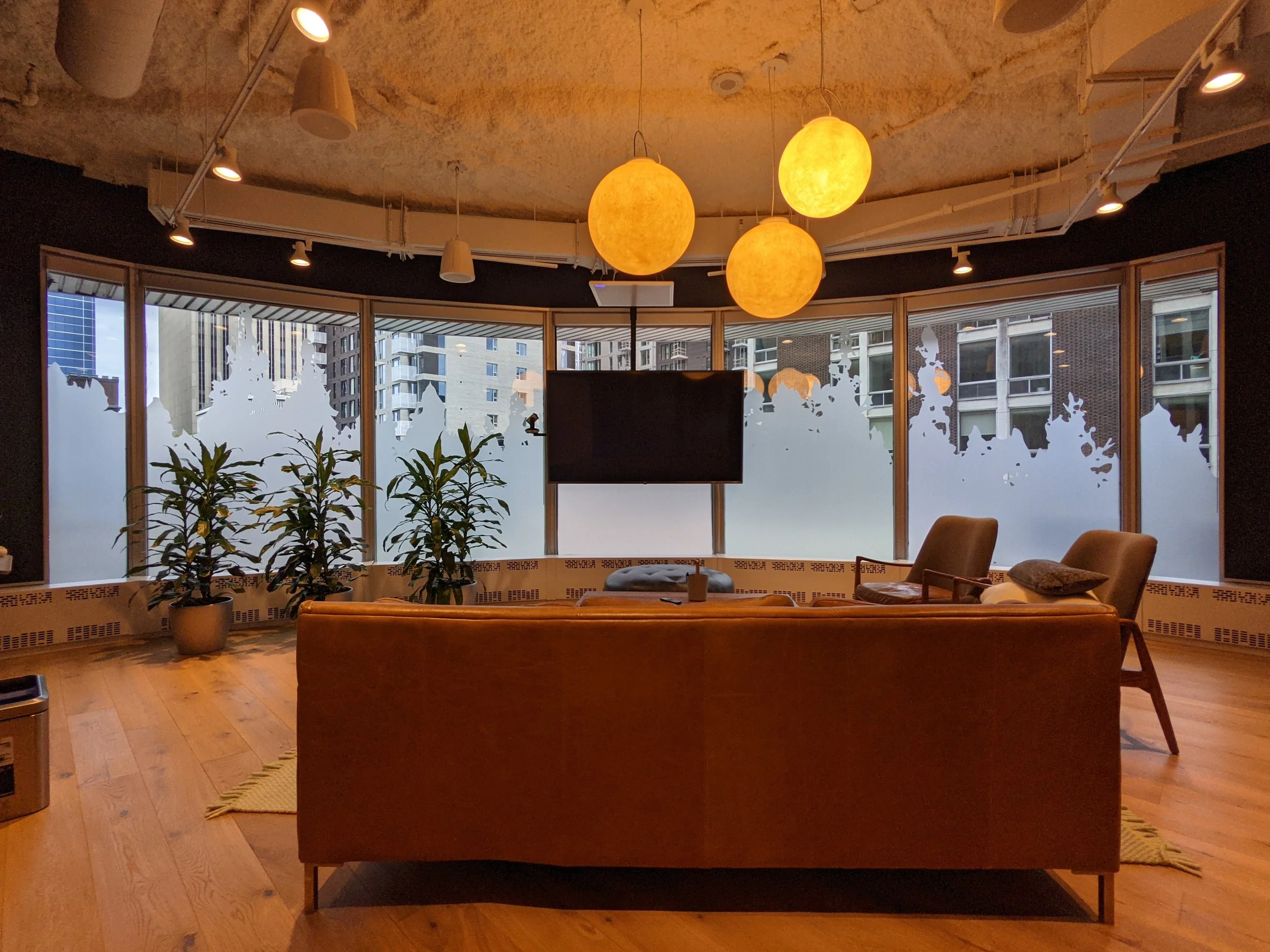 Interior of a room with a curved glass window wall, wooden flooring, and modern lighting fixtures including three glowing spherical ceiling lights and track-mounted spotlights. There are potted plants and furniture visible.