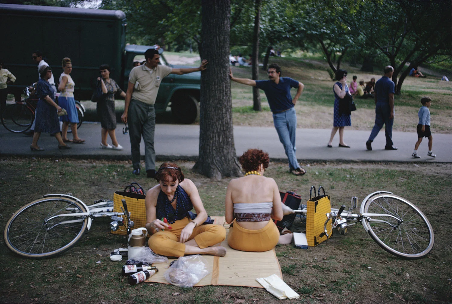 Two women in mustard-yellow trousers sit on a straw mat in a park with bicycles laid flat on the grass either side of them like wings; behind on the path, two men each brace a hand against a tree trunk in near-identical poses, with a truck, a boy and scattered pedestrians beyond