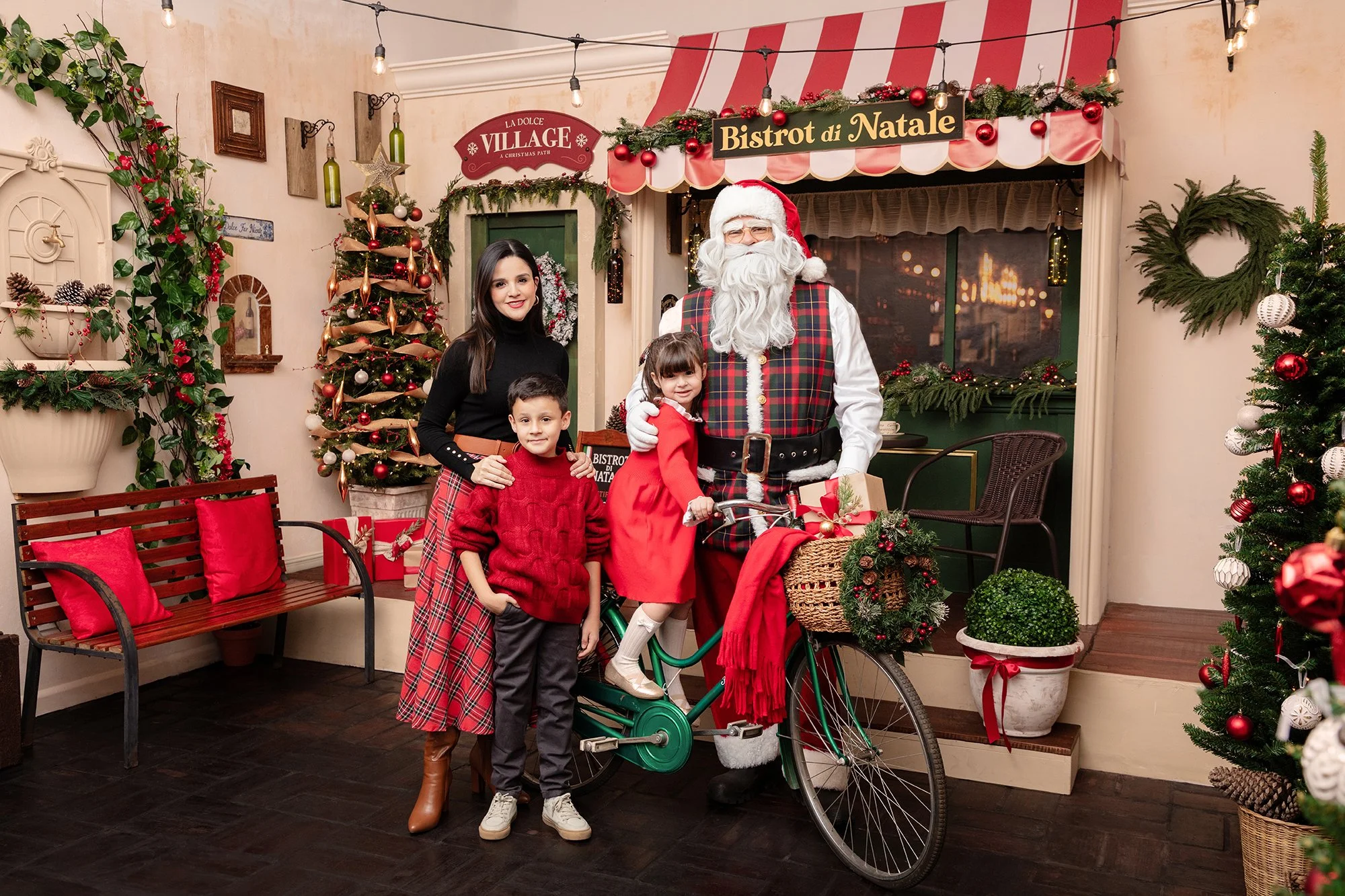 Niña y niño con madre con Santa Clus en celebración navideña en un espacio decorado con árbol de Navidad, guirnaldas, lazo dorado, bolas rojas y blancas, y luces navideñas, en un ambiente festivo y decorativo. Sesión fotos navideña ciudad Guatemala.