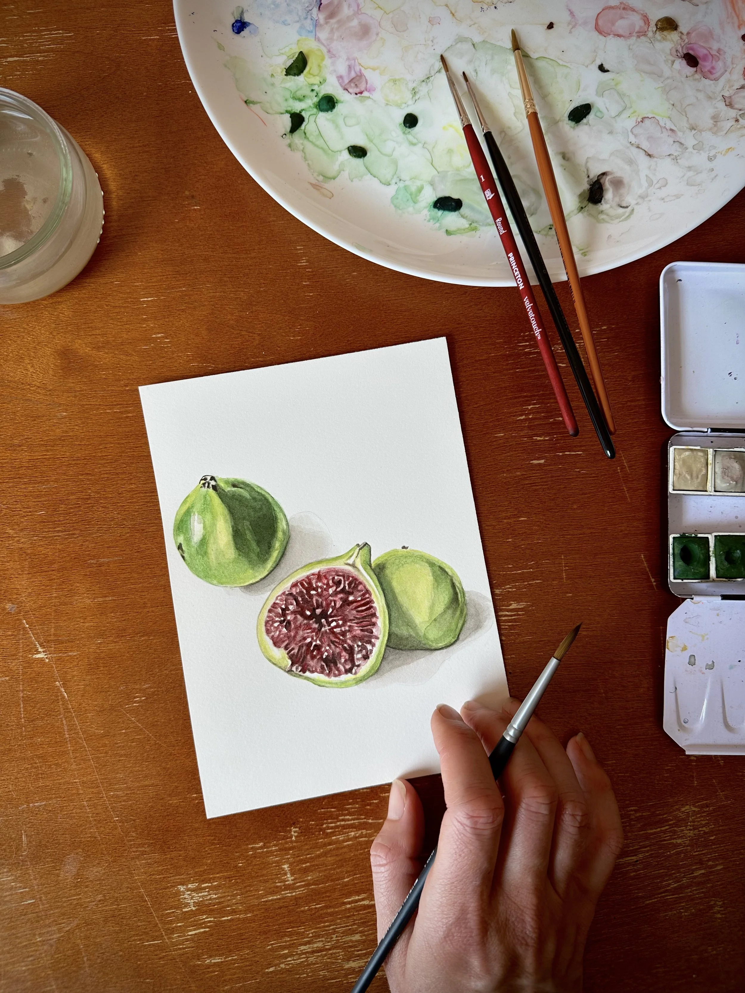 A person painting a realistic watercolor illustration of three green figs, one of which is cut in half showing its red interior, on a white sheet of paper on a wooden table.