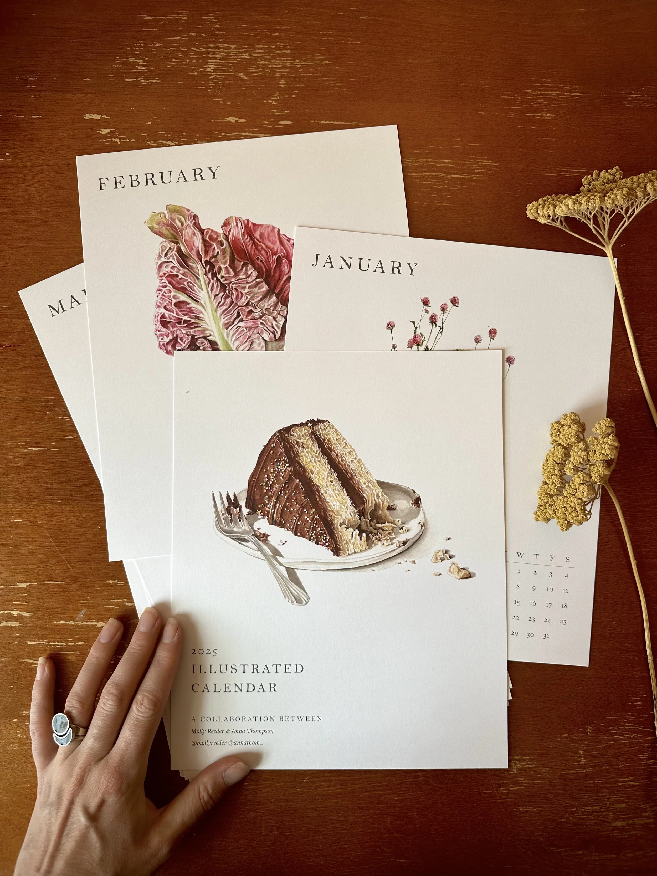 A person holding a 2025 illustrated calendar with floral artwork on a wooden table, along with additional calendar pages visible, and some dried flowers on the table.