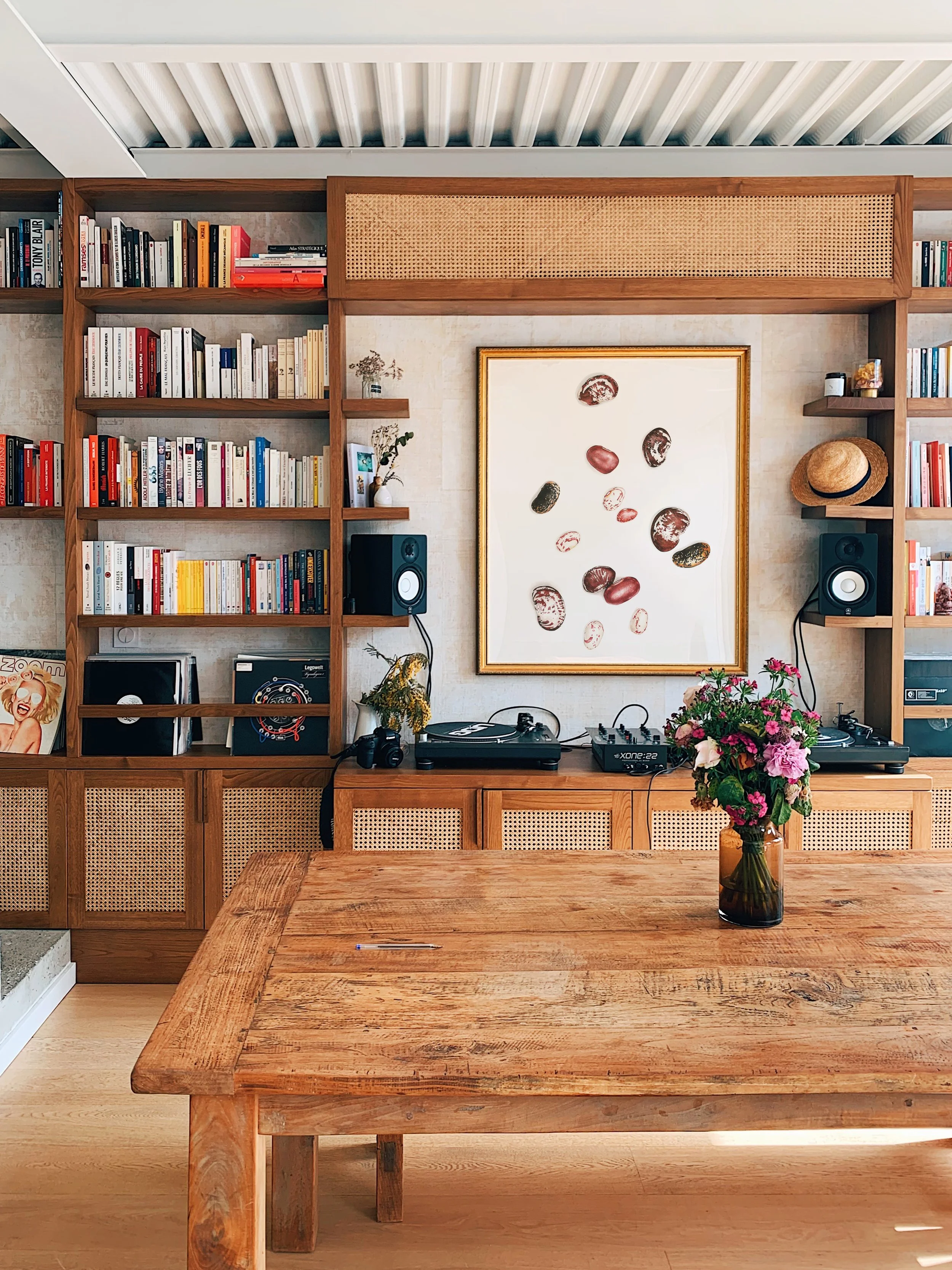 A wooden dining table with a vase of pink and purple flowers in the foreground. In the background, a wall with built-in shelves filled with books, a framed art piece with beans, and music equipment including turntables and speakers.