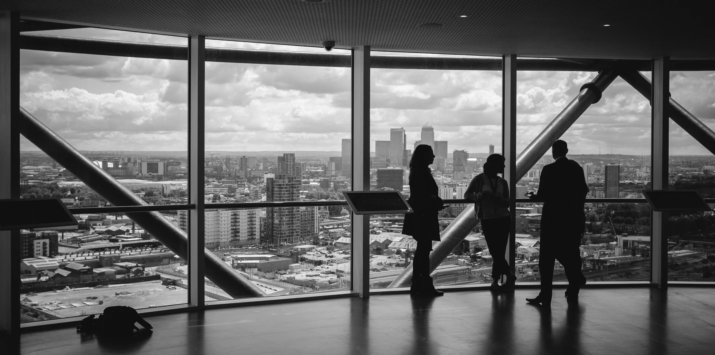 Silhouettes of four people standing and talking inside a high-rise building with large windows, overlooking a city skyline with tall buildings and a partly cloudy sky.
