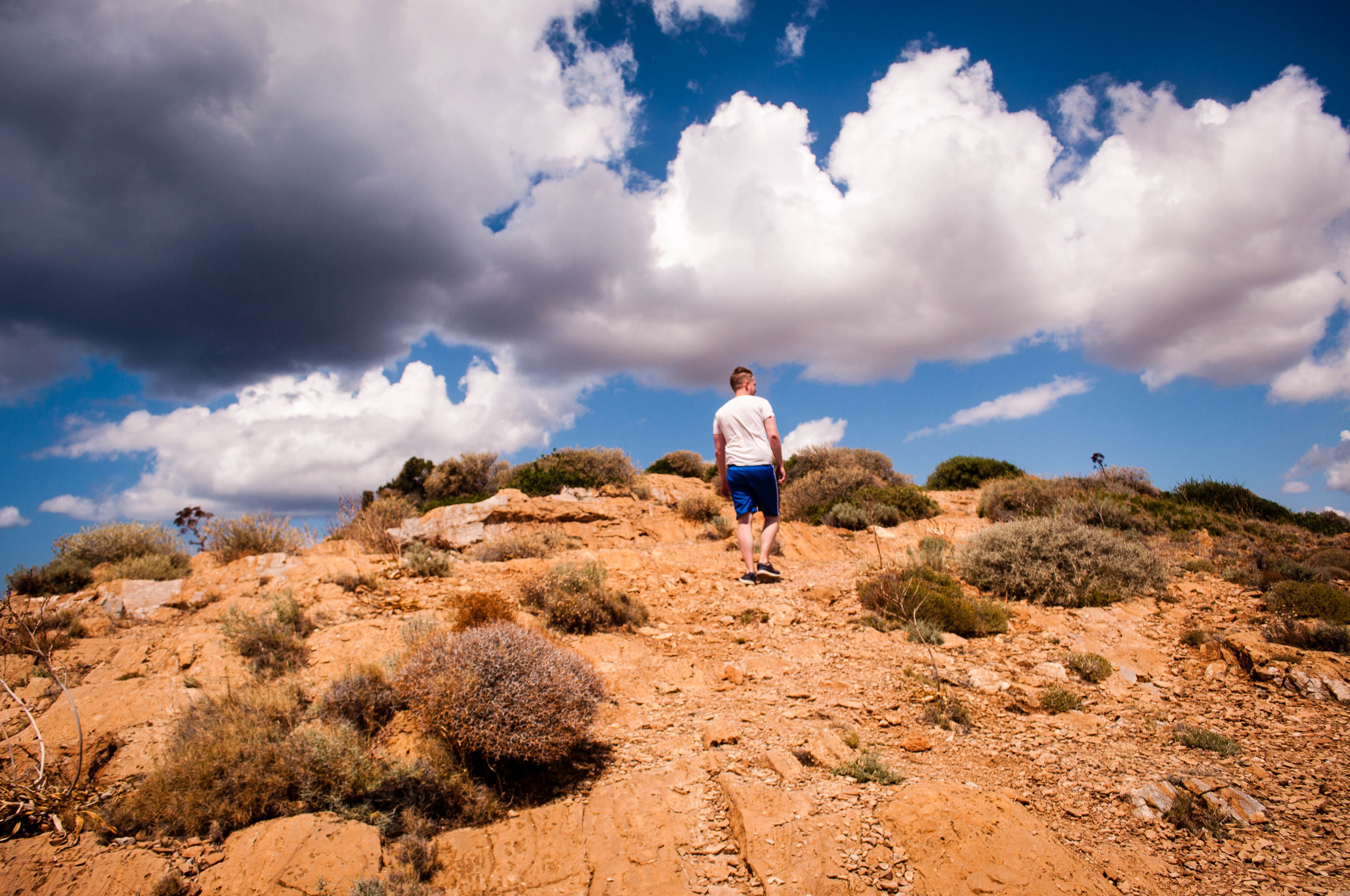 Blue sky with clouds with a white man walking near a cliff on a terracotta ground  in Sounion (Greece)  