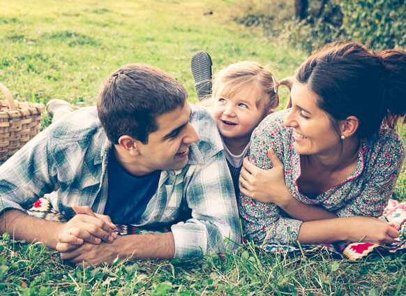 Smiling parents lying on grass outdoors with their baby between them, enjoying family time together on a sunny day