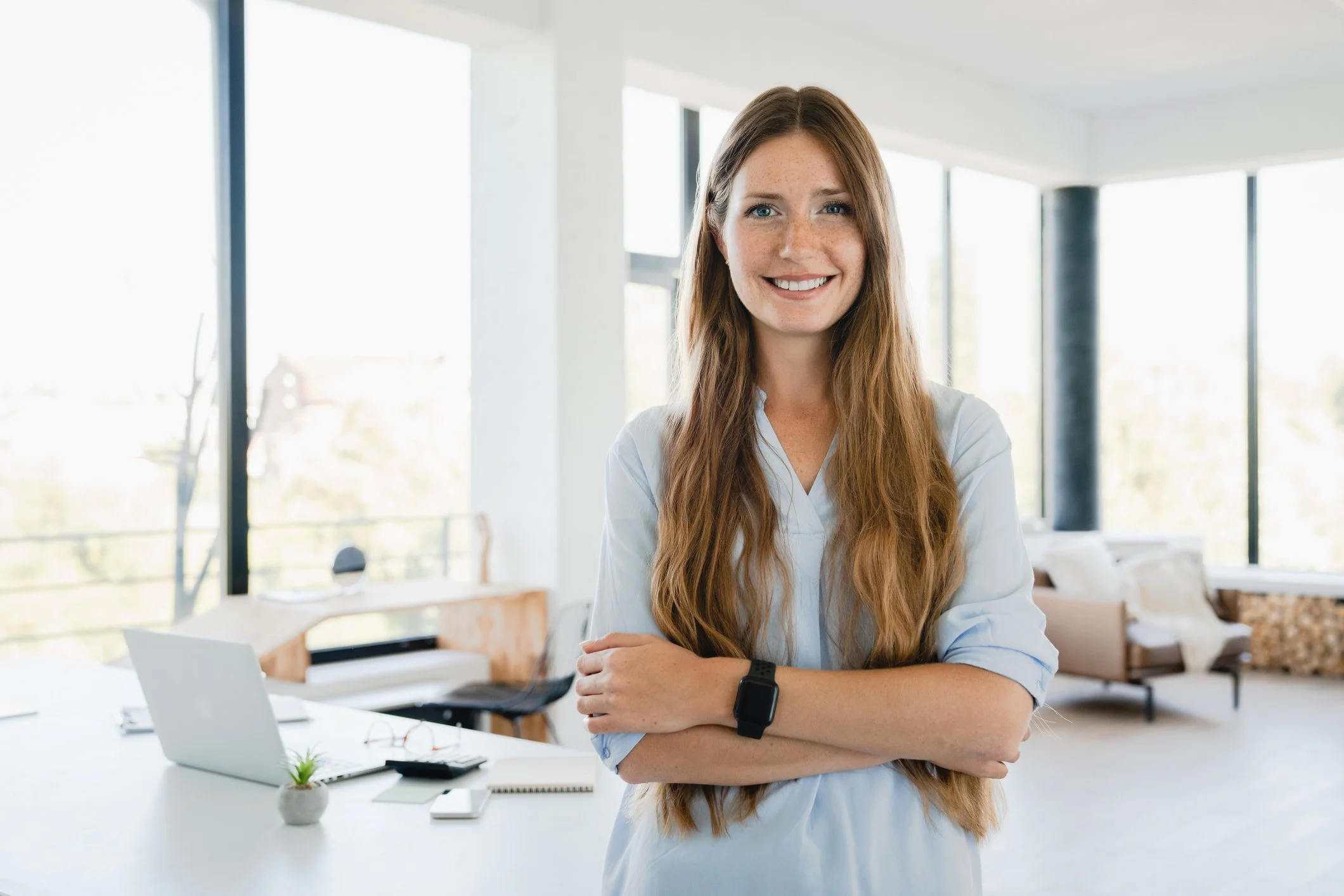 Smiling professional woman standing with arms crossed in a bright modern office, conveying confidence and approachability.