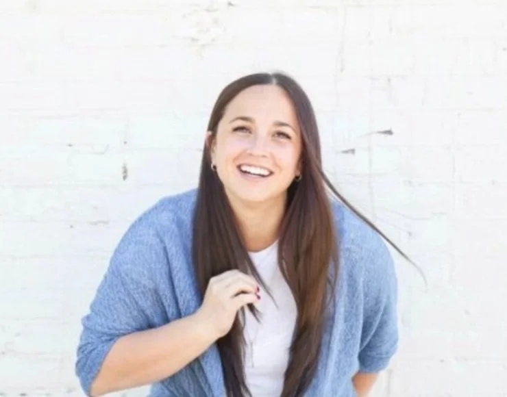 Smiling owner Kate standing against a white brick wall in a natural light portrait