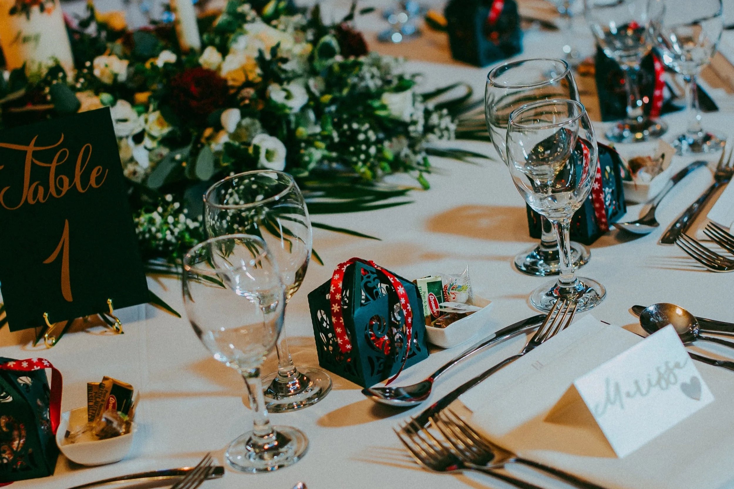 A decorated banquet table set for a celebration with wine glasses, silverware, and holiday-themed decorations, including small gift boxes and a floral centerpiece.
