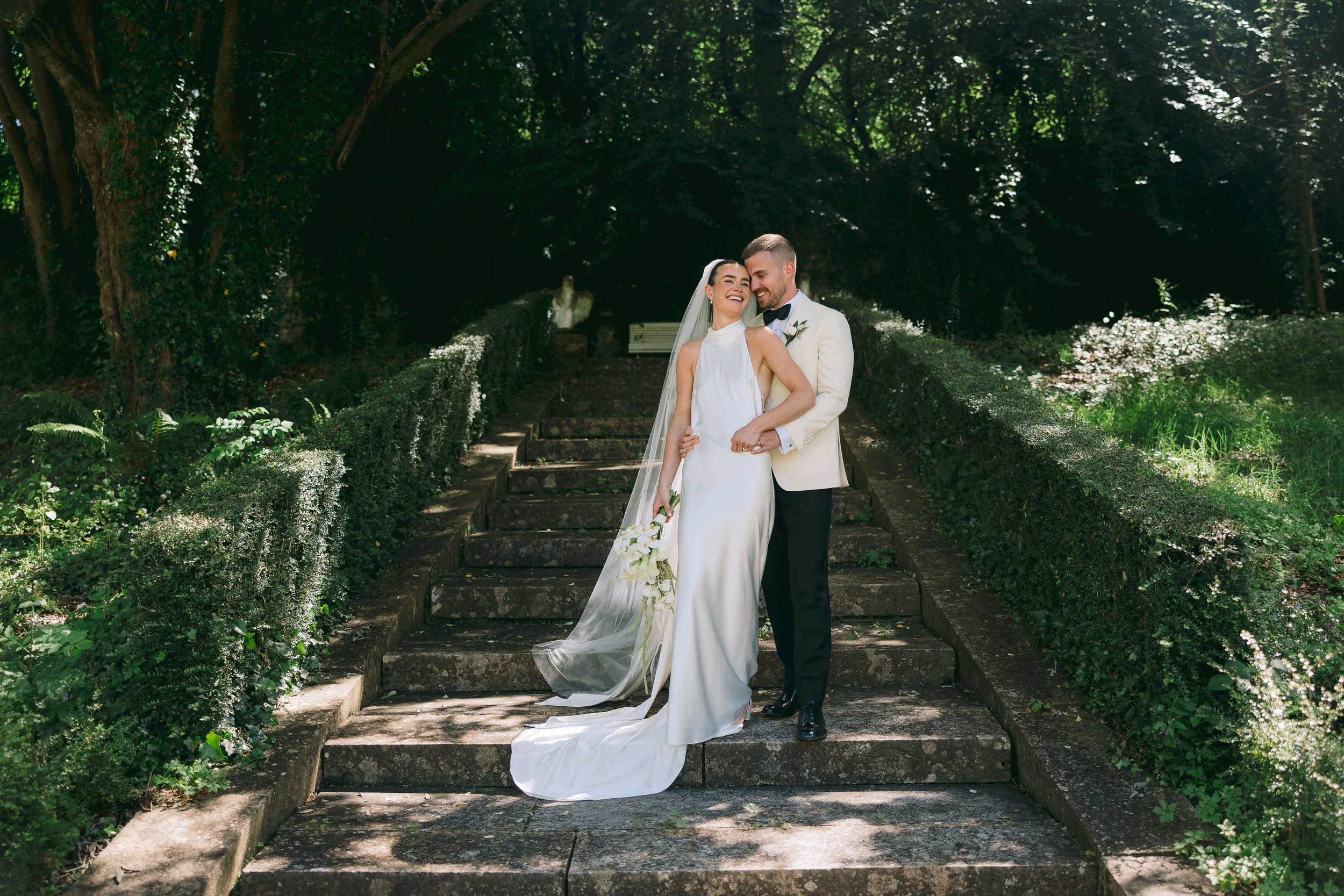A newlywed couple smiling and embracing on stone steps in a lush green garden, with tall trees and white flowers surrounding them.