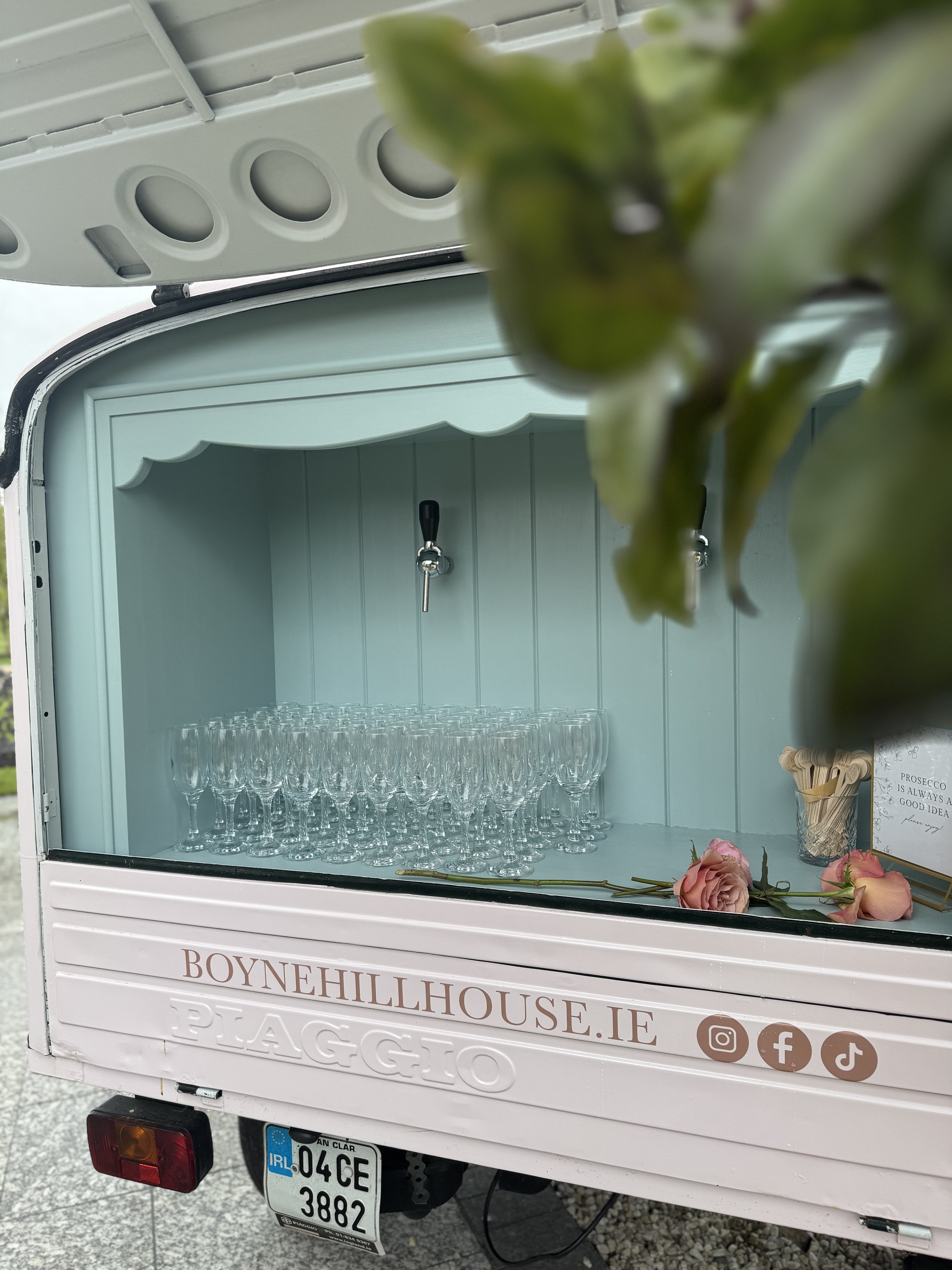 A mobile bar cart with glasses and flowers, possibly for serving drinks at an event or gathering, parked outdoors.