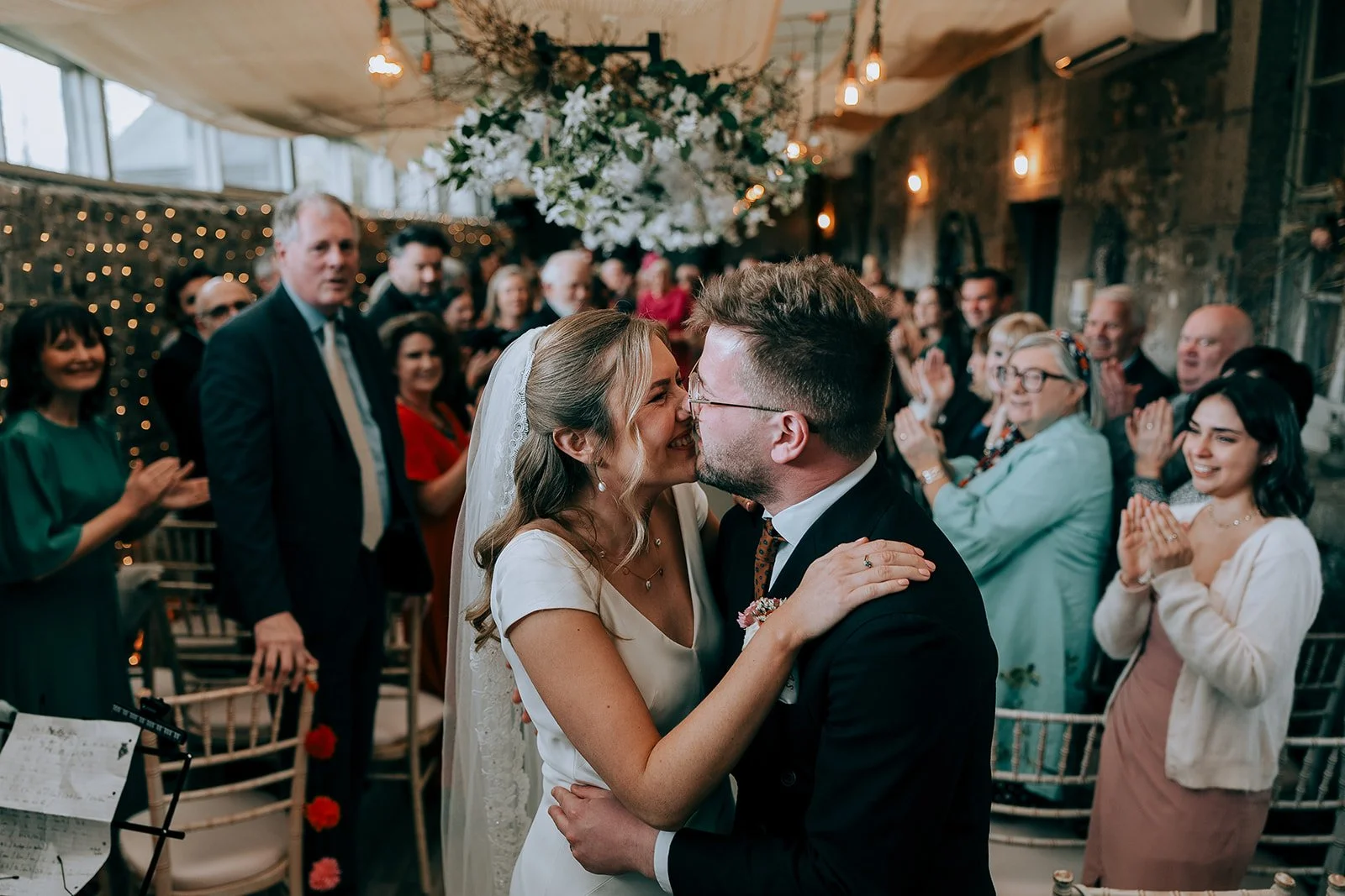 A couple sharing a kiss during a wedding ceremony in a decorated venue surrounded by happy friends and family.