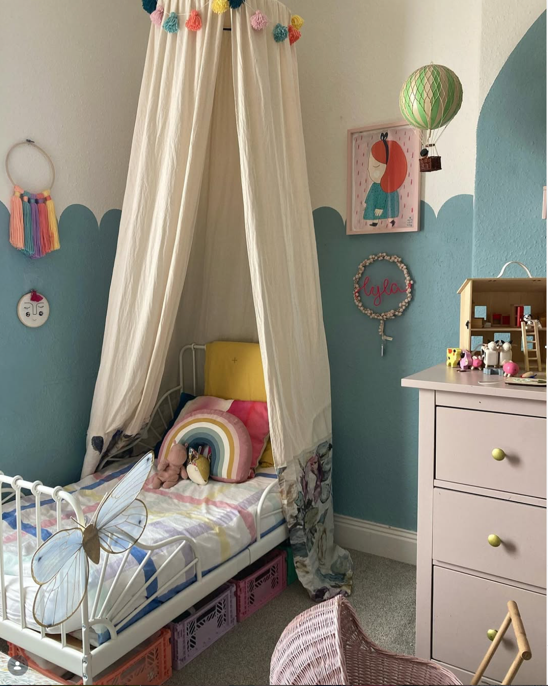 Cosy children's bedroom with a cream fabric canopy over a white bed, turquoise scalloped walls, colourful cushions and a personalised Lyla light wreath.