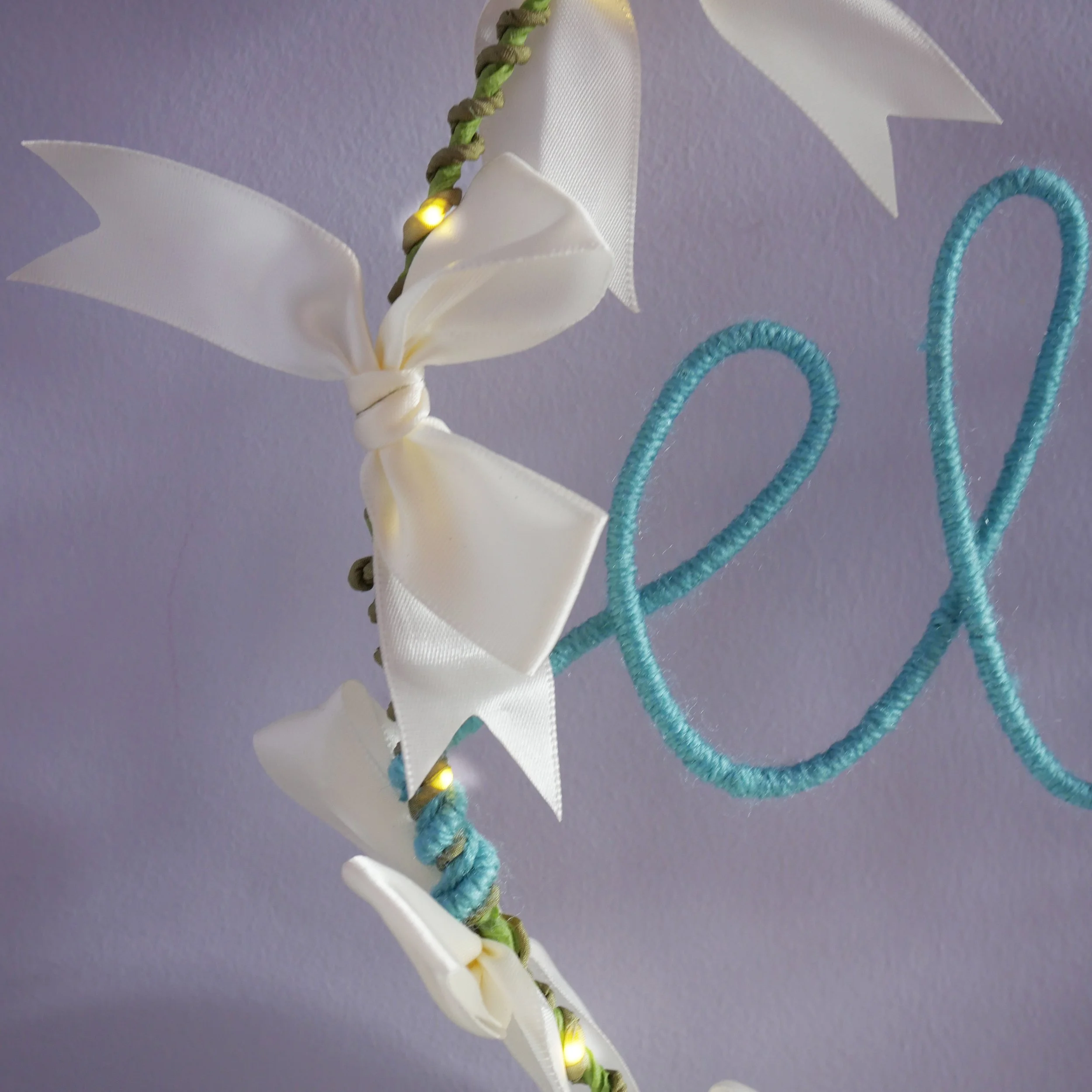 lose-up of ivory satin bows, olive green wrapped hoop and duck egg blue yarn lettering with warm white LEDs.  Copy