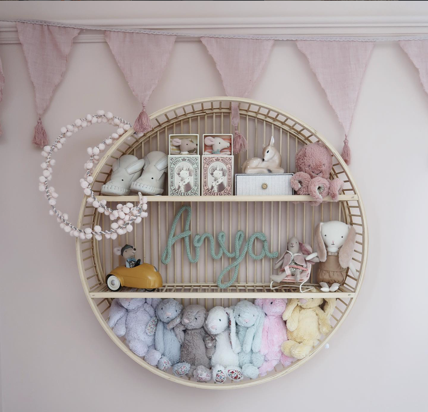 Round rattan nursery shelf displaying soft toys, keepsakes and a personalised Anya name sign, with a white pom pom fairy light moon beside it.