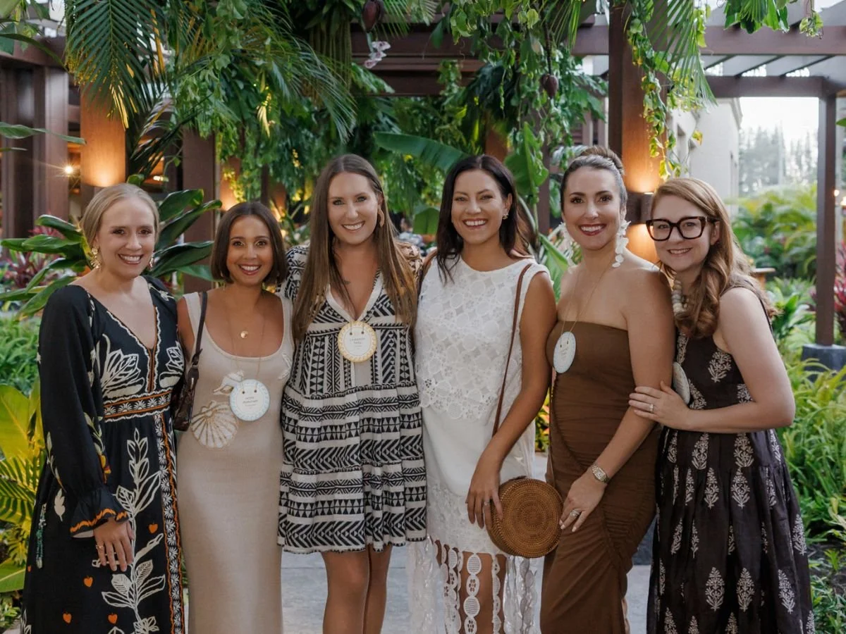 Group of six women posed for photo in front of a backdrop of tropical ferns