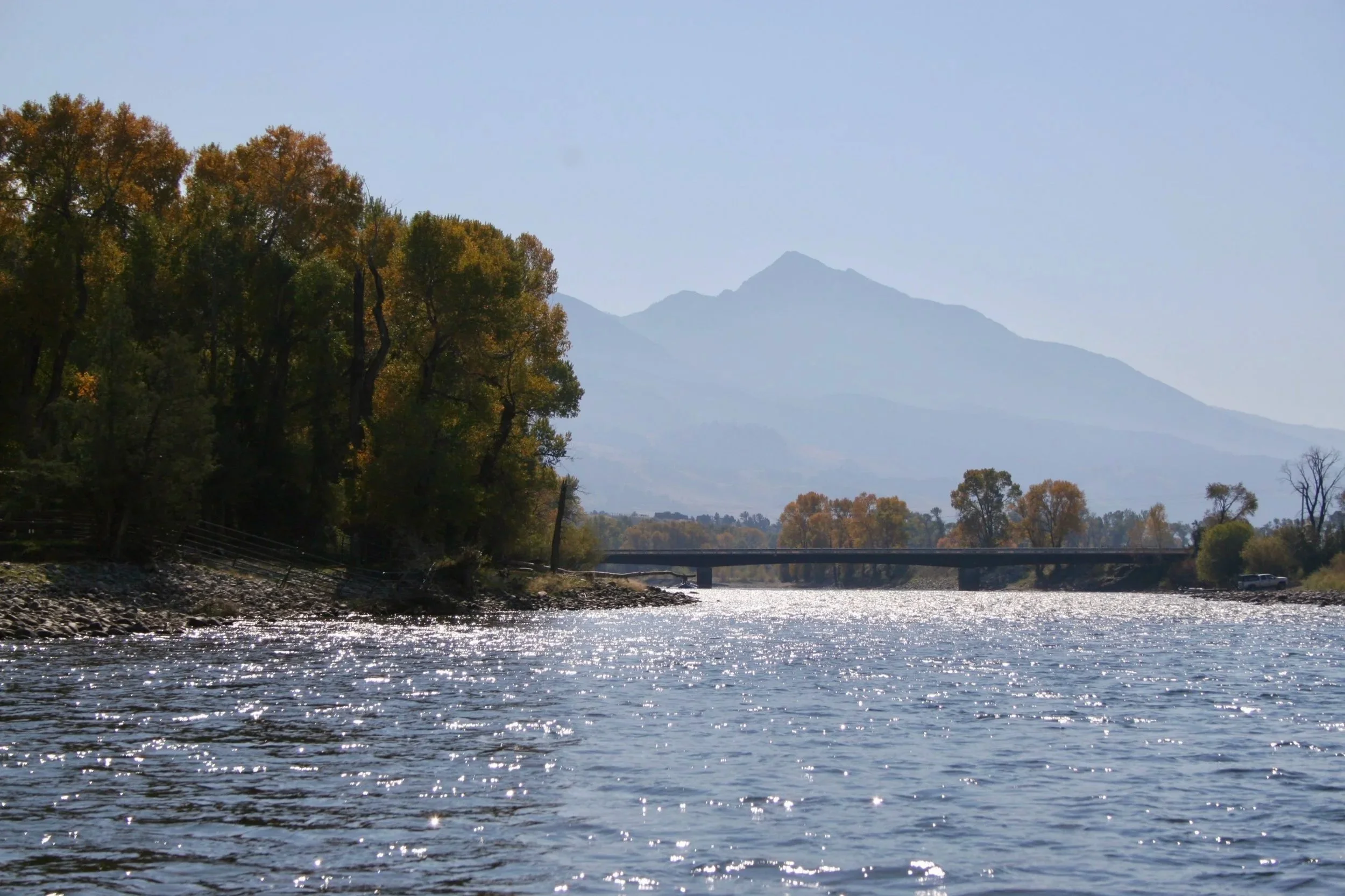 A scenic view from the water looking upriver toward the historic Pine Creek Bridge on the Yellowstone River, with the jagged peaks of the Absaroka Range in the background during a guided float trip in Paradise Valley.