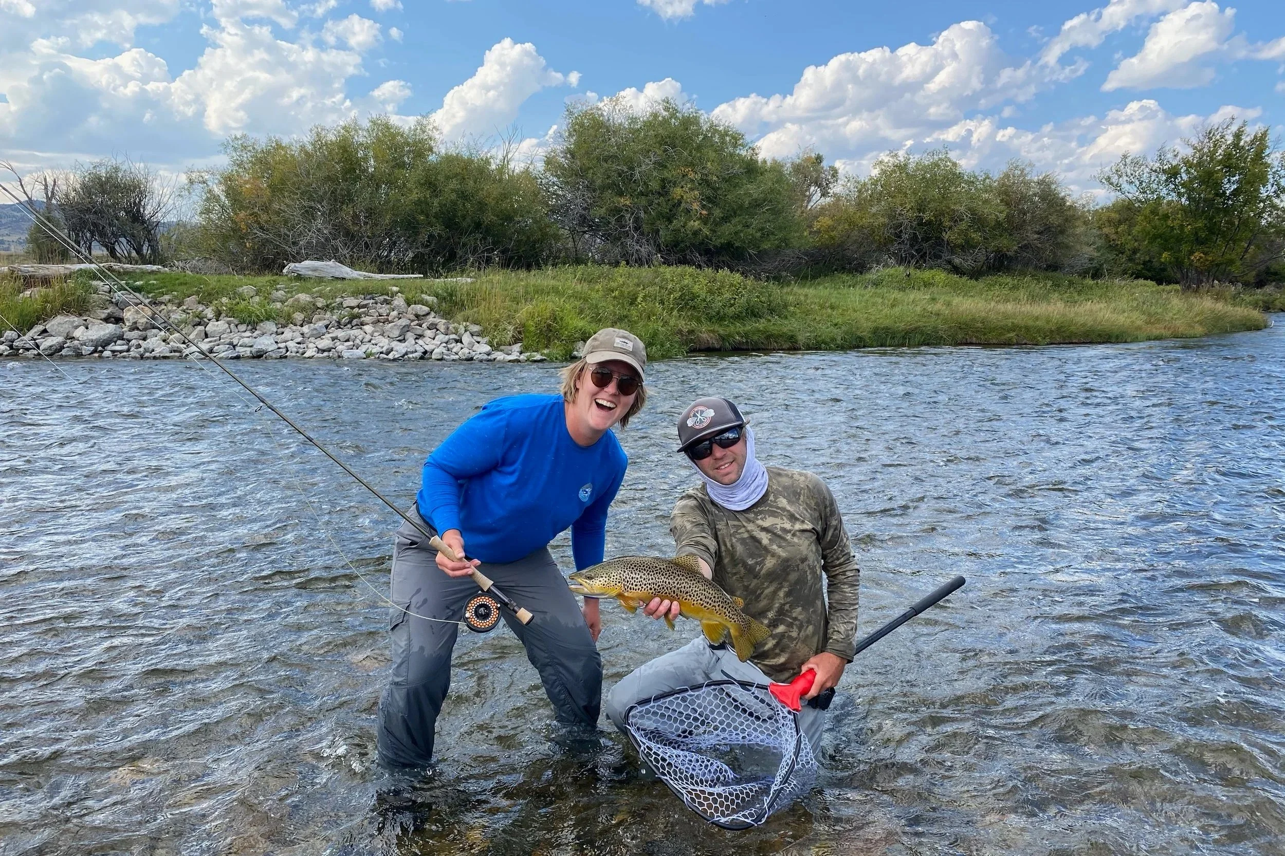 A guide kneeling with a net and a Madison River brown trout while a smiling client leans in during a summer walk-and-wade fishing trip near Bozeman, Montana with Flyshot Outfitters.