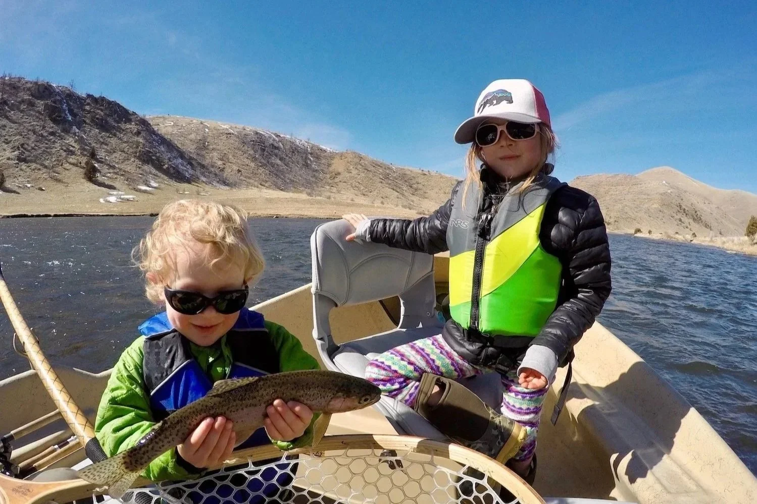 A young boy smiling and holding a rainbow trout while his sister looks on from a drift boat during a safe, family-friendly fly fishing trip on the Madison River with Flyshot Outfitters.