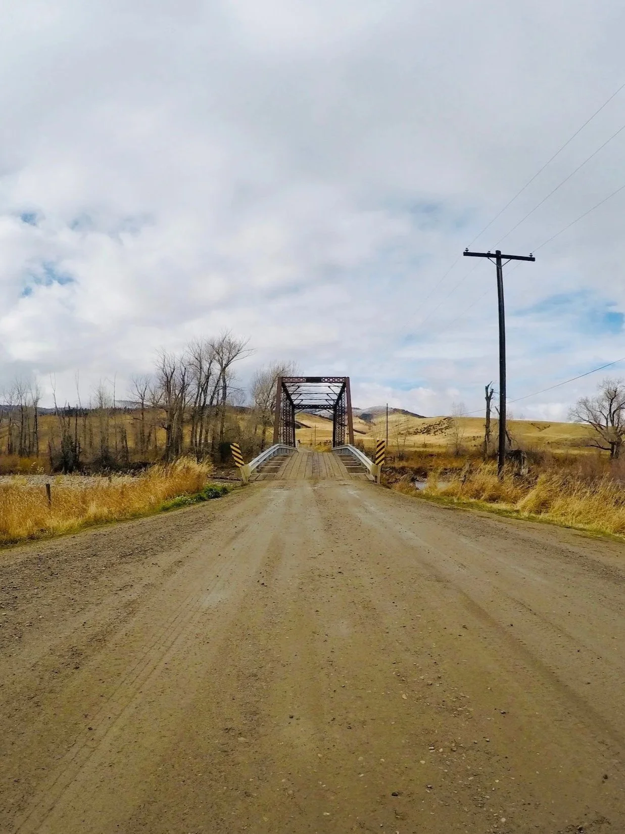 A historic pre-flood view of the Williams Bridge on the Gallatin River near Gallatin Gateway, seen from the road with the Gallatin Mountain Range in the background before the bridge was moved off its pilings in the 2022 flood year.